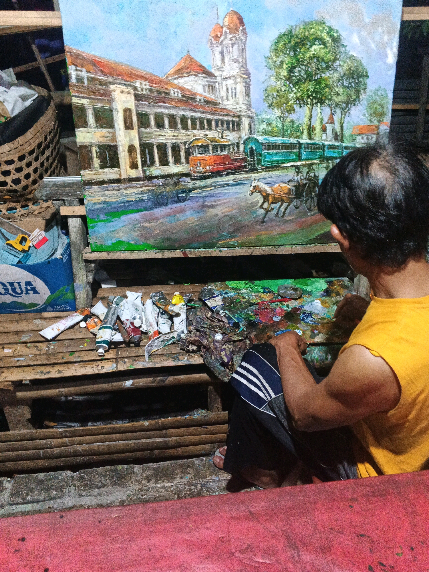 The image depicts a man in a yellow t-shirt painting an oil on canvas artwork that appears to be a street scene in Ungaran, Indonesia. The painting features a colonial-era building with a prominent tower, a red tram, a turquoise train, and a horse-drawn carriage with two figures. The artist is seated on a wooden surface, working on the painting. In the foreground, on a wooden plank, are tubes of oil paint, a palette with mixed colors, and various painting tools. To the left of the artist, there is a woven basket filled with white material and a blue plastic container of "AQUA" bottled water. The background shows the artist's workspace, with shelves holding more art supplies and a partially visible painting. The overall scene conveys an artistic and somewhat cluttered environment.