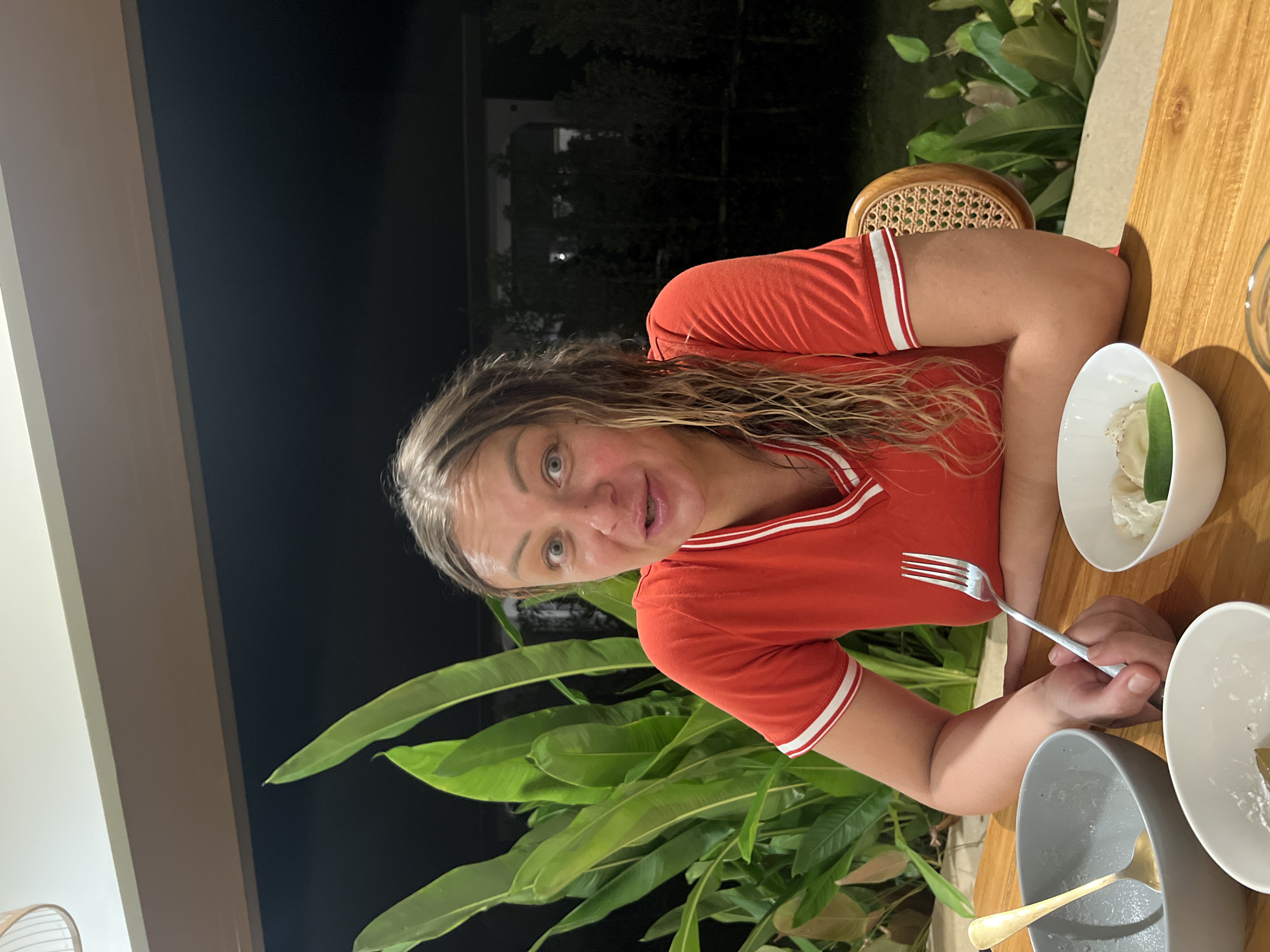 A medium shot shows a woman sitting at a wooden table outdoors at night, with a fork in her hand. She has long, wet, blonde hair and is wearing a red t-shirt with white trim. Her eyes are wide open, and her mouth is slightly ajar as if she is speaking. In front of her are three bowls, two empty and one with food. Behind her, large green leaves are visible against a dark background, suggesting a tropical setting. The overall scene appears to be a restaurant or dining area in Koh Phangan, Thailand, given the environment and time of day.