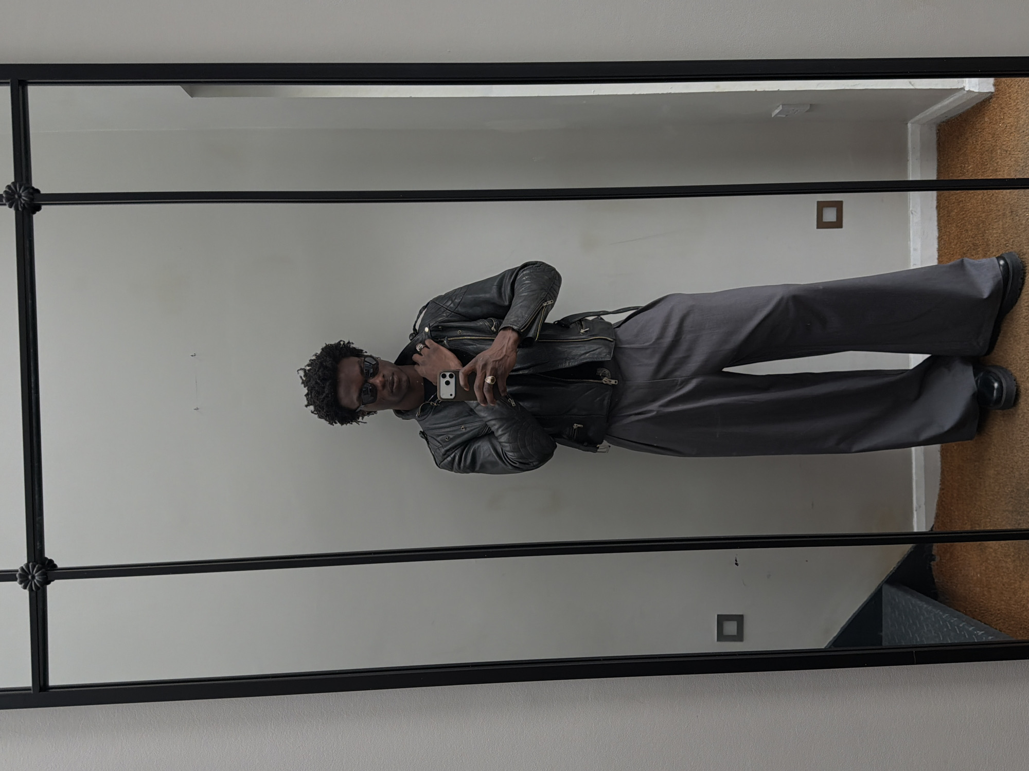 A man stands in front of a mirror, facing the camera. He is wearing sunglasses, a black leather jacket, gray pants, and black shoes. He holds a smartphone in his right hand, presumably taking a photo. The mirror has a black frame with decorative elements. The background consists of a white wall with two square electrical outlets, a section of carpet, and a staircase. While not confirmed, it is a plausible setting for a flat or townhouse in a city like London, United Kingdom.