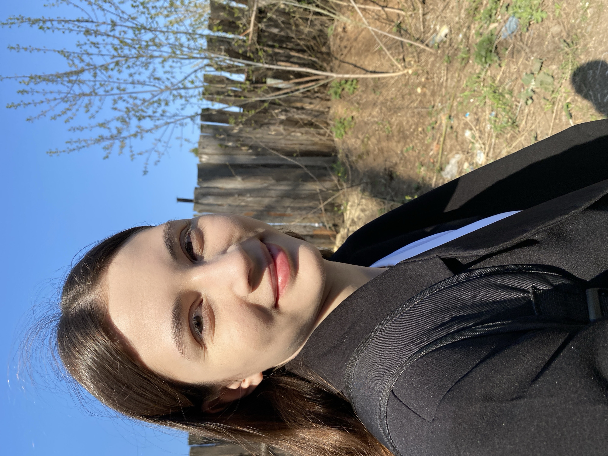A medium-shot, candid, outdoor photograph captures a young woman in sunlight against a backdrop featuring a dilapidated wooden fence, sparse vegetation, and a tree. The woman, positioned at an angle relative to the camera, has fair skin, dark brown hair, and light makeup. She is wearing a black jacket over a white garment, with part of a black backpack strap visible on her shoulder. The background, including the fence and vegetation, suggests a possibly neglected or rural edge of an urban area. The blue sky is visible above the fence and tree branches. Given the prompt's context, the location could potentially be a residential or peripheral area within Votkinsk, Russia.