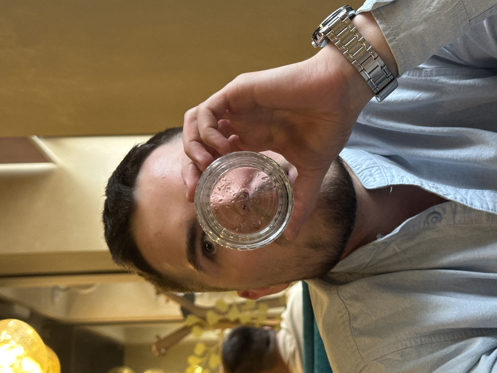 A person holds a glass containing a brownish-pink substance up to their eye. The person has dark hair, a beard, and is wearing a silver watch and a light-blue button-down shirt. A second person is visible in the background, out of focus. A blurry background is visible. It is not possible to confirm that the location is in Perm, Russia, or to state any interaction between people.