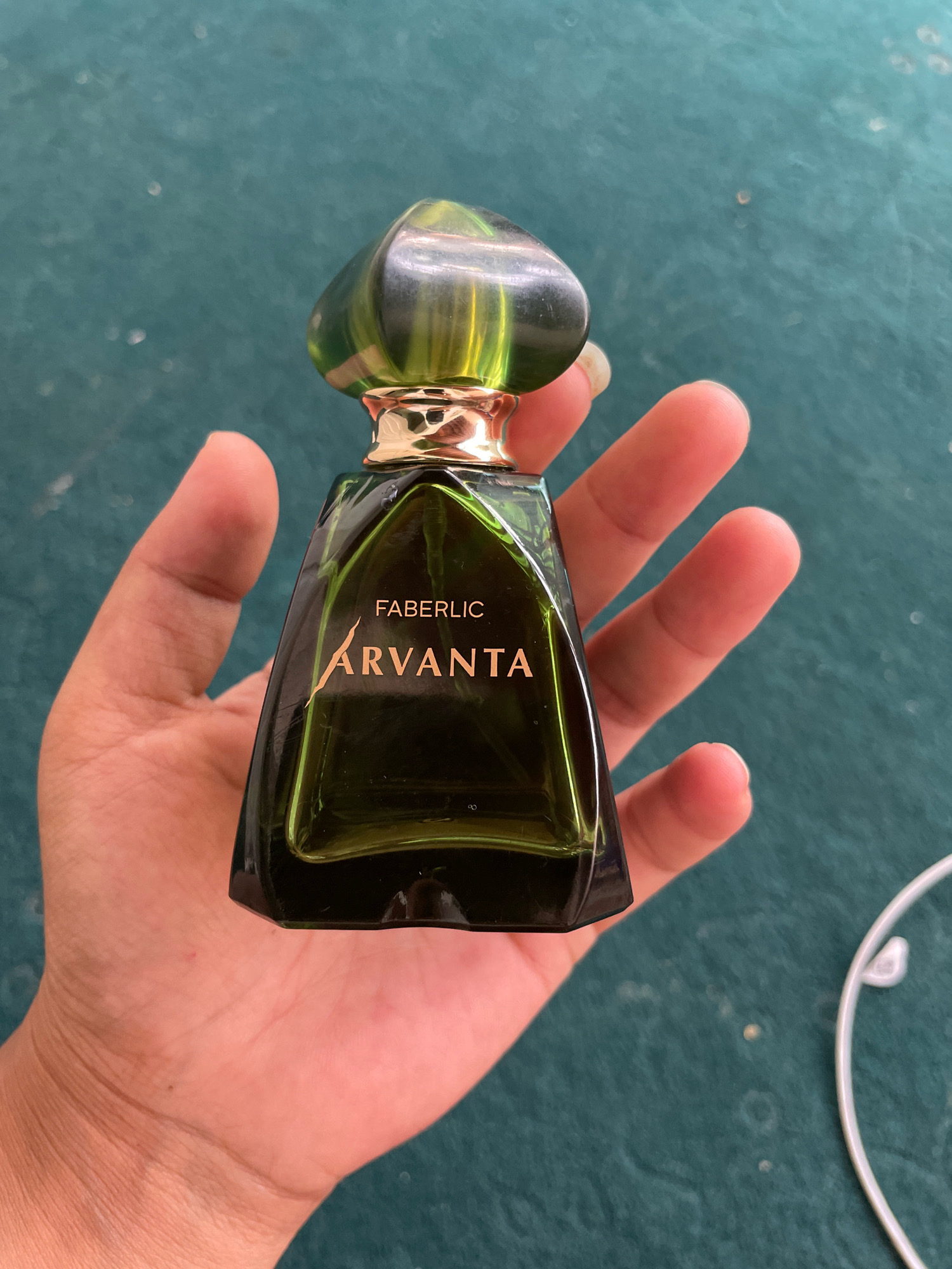 A hand holds a green glass perfume bottle with a gold cap and gold lettering. The lettering on the bottle reads "FABERLIC ARVANTA". The background is a dark teal textured surface. No location in the city of Чекелик, Kyrgyzstan is visible or implied.