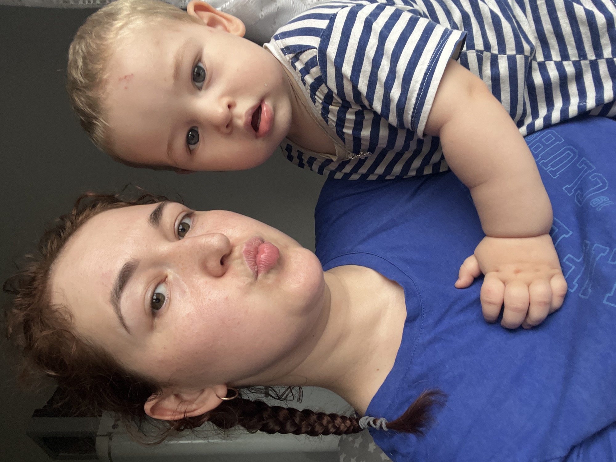 This is a selfie of a woman and a baby. The woman has reddish-brown hair in a braid and is wearing a blue t-shirt. She is puckering her lips and holding the baby in her arms. The baby has blonde hair and is wearing a blue and white striped shirt. The background is out of focus and appears to be indoors, with some gray wall and a glimpse of a white curtain or fabric. There is no indication of the location being Chelyabinsk, Russia.