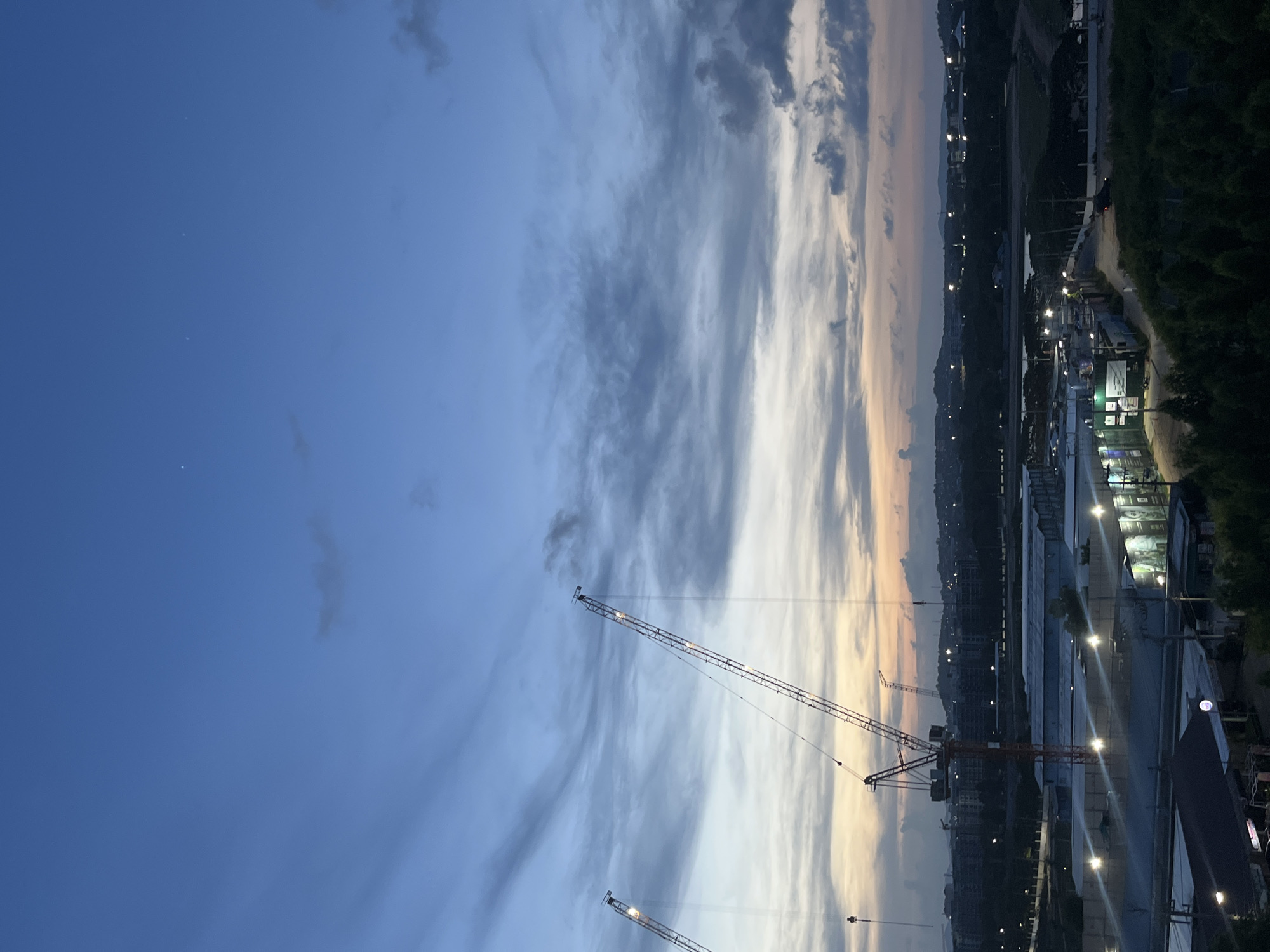 The image shows an evening scene in Pattaya City, Thailand. The sky is a mix of blue and grey clouds with hints of orange near the horizon, indicating a sunset or twilight. In the foreground, there are multiple construction cranes, suggesting active development or building sites. In the background, there are city lights, buildings, and greenery, possibly indicating residential or commercial areas. The perspective is from an elevated position.