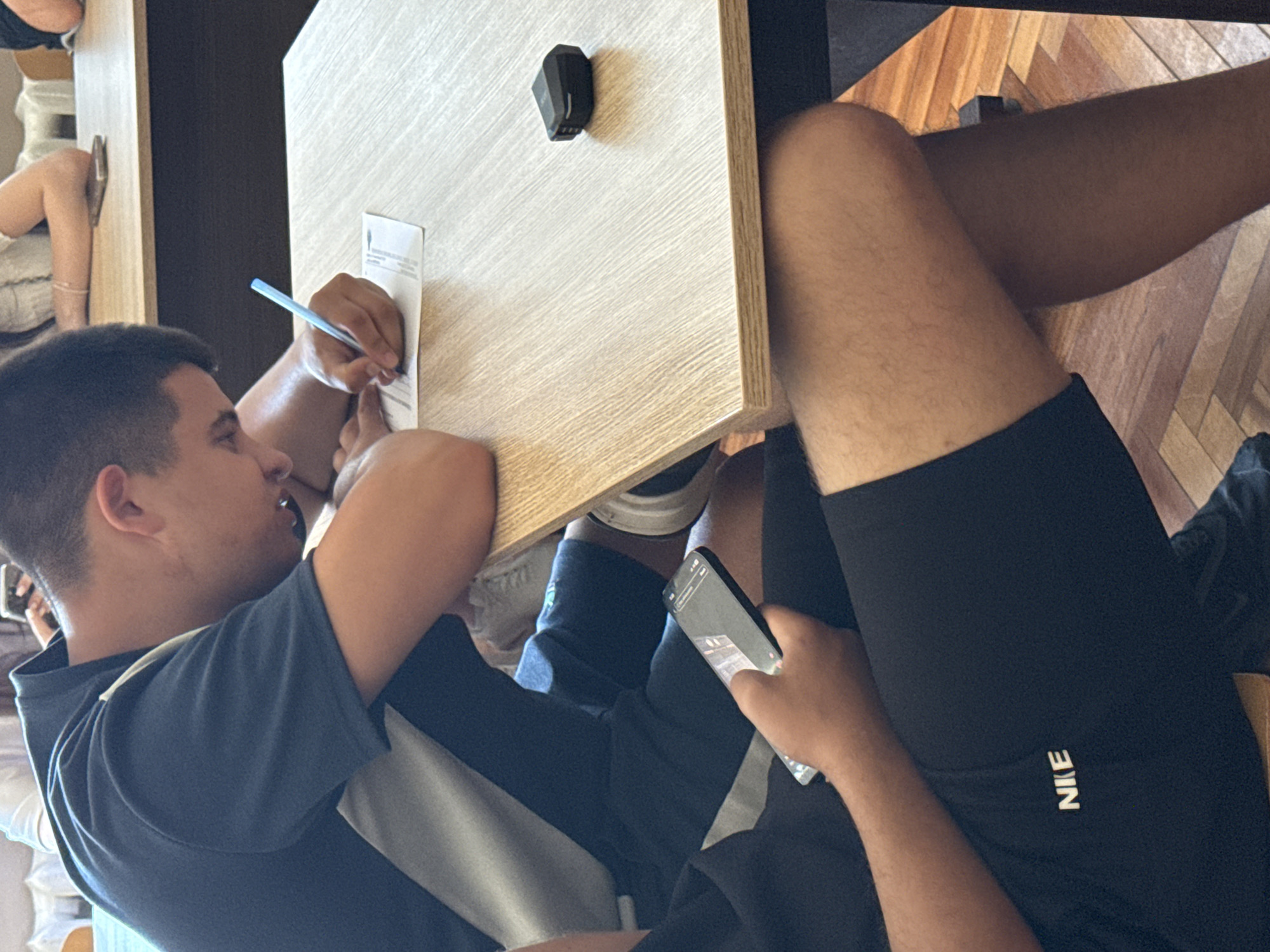 A man with short dark hair is seated at a light-colored table, writing on a piece of paper with a blue pen. He is wearing a blue t-shirt with white stripes. Another person, wearing black Nike shorts, sits to his right, holding a cell phone in their hand. A small black object is on the table next to the paper. The background suggests an indoor setting with wood flooring. The location and city are not discernible in the image.
