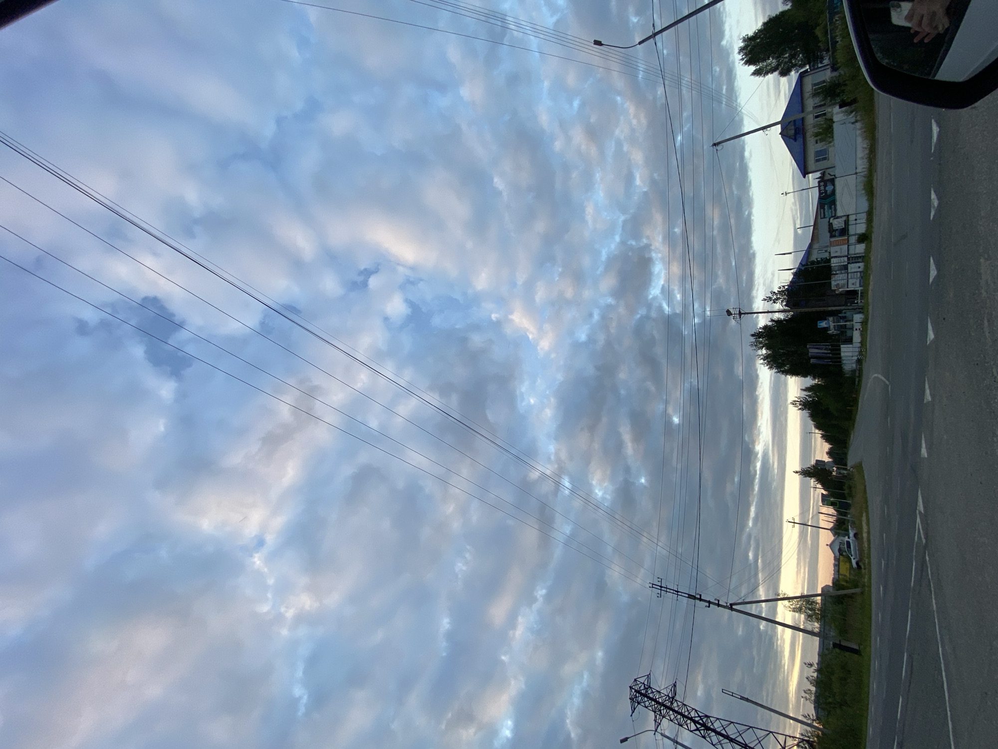 The image captures a view of a road intersection in Nyagan, Russia, under a cloudy sky. The sky is filled with a mix of light and dark grey clouds, with hints of blue peeking through in some areas. Multiple power lines stretch across the frame, supported by utility poles and a large metal pylon on the left.
The foreground shows a paved road with white markings indicating lanes and a pedestrian crossing. To the left of the road, there is a grassy embankment with bushes and trees. A silver pickup truck is parked near the embankment. Further ahead, several buildings with blue roofs are visible, along with fences and signage. Trees line the sides of the road in the distance. The overall atmosphere suggests an overcast day, possibly during the early morning or late evening given the lighting. The scene is devoid of any people actively engaged in the environment, although a hand holding a phone is visible in the rearview mirror of a vehicle, indicating the photo was taken from inside a car.