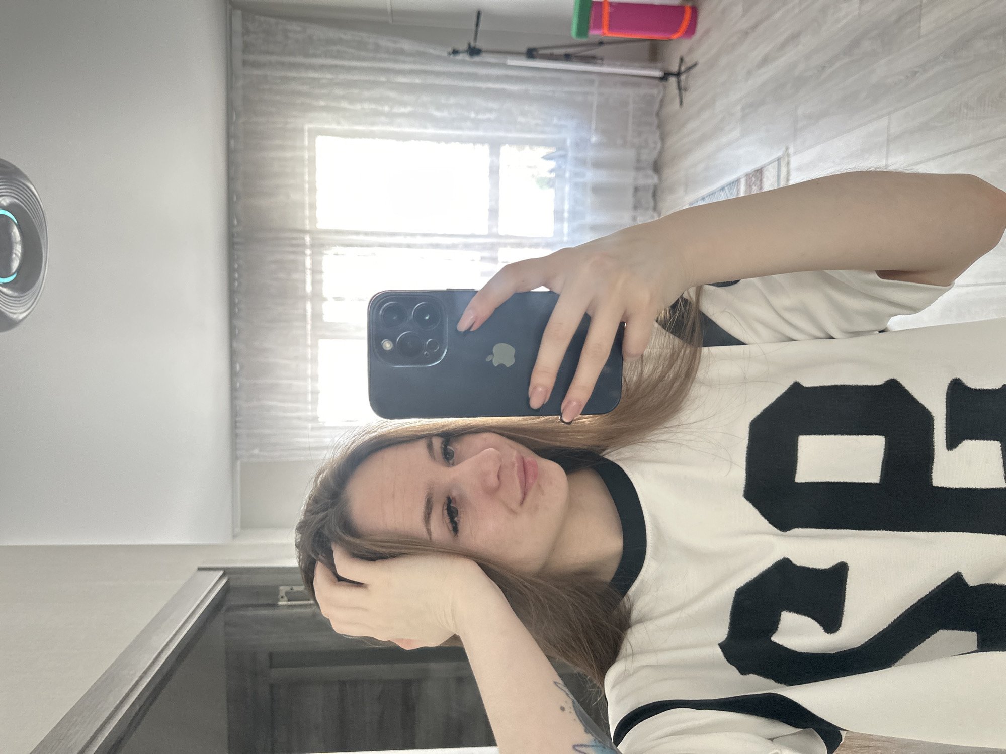 An image, rotated 90 degrees counter-clockwise, captures a young woman taking a mirror selfie indoors during the daytime. The woman, with long brown hair, looks directly at the camera with a neutral to slightly smiling expression. Her left hand rests near her head, and a tattoo is partially visible on her left forearm. She is wearing a white top with black trim around the neck and sleeves, and a large black number "89" is prominently displayed on the front.
Her right hand holds a dark-colored smartphone, likely an iPhone due to its three camera lenses and visible Apple logo on the back. The background shows the interior of a room, brightly illuminated by natural light entering through a large window covered by sheer white curtains or blinds. On the white wall to the left, a circular, silver-toned device with a glowing blue light is mounted. Reflected behind the window, a light stand or tripod is visible, topped with colorful items in pink and green. The floor appears to be made of light-colored wooden planks or laminate, and a dark-colored cabinet or piece of furniture is visible in the lower left foreground. The bright lighting indicates it is daytime.