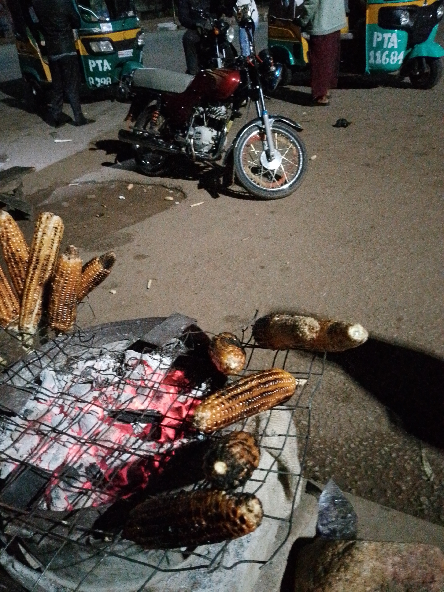 In Bukuru, Nigeria, an outdoor scene at night depicts food preparation and transportation. A motorcycle, a common mode of transport in the city, is parked on a paved surface. In the foreground, corn is being grilled over hot coals, with several cobs already on the grill rack and others waiting to be cooked. Behind the grilling station, a three-wheeled auto-rickshaw, also known as a Keke Napep, is visible. These vehicles are a popular form of public transportation in Nigerian cities. The image captures a slice of everyday life, showing a common method of food preparation and the prevalent modes of transportation in Bukuru.