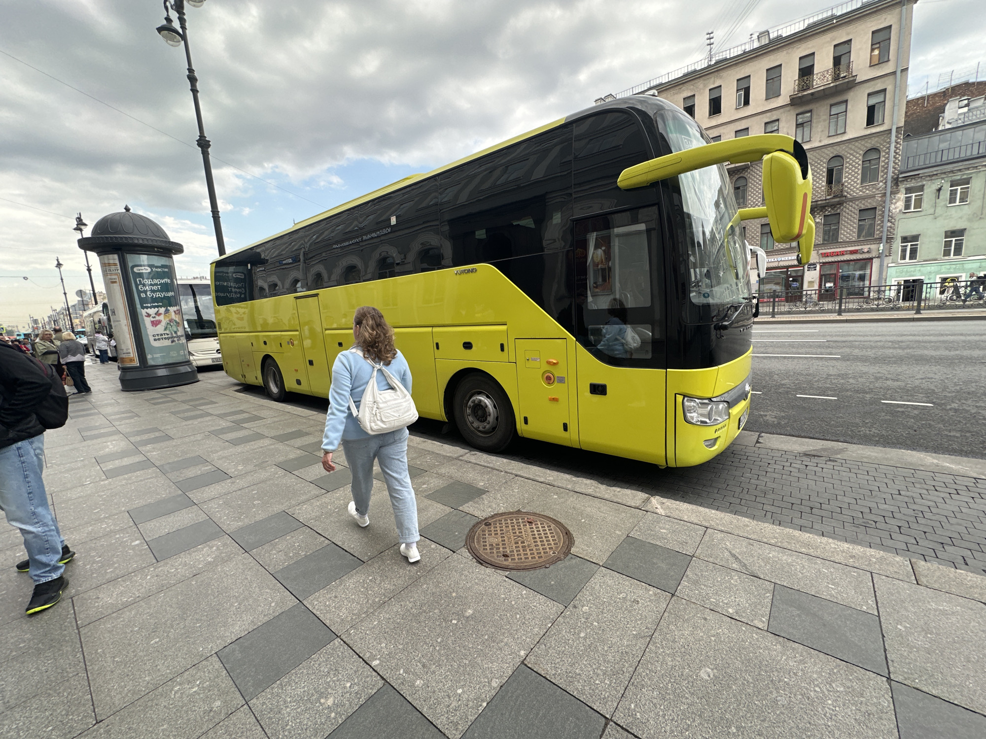 The image shows a yellow XUYONG bus parked on a street in Saint Petersburg, Russia. A woman with blonde hair is walking on the sidewalk next to the bus, carrying a white purse. A man in jeans and a black jacket is walking in the foreground, partially cropped out of the image. In the background, there is a kiosk with a poster on it. A tall building is visible behind the bus. The sky is cloudy.