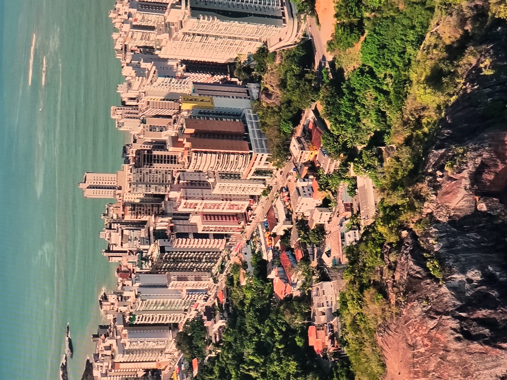 This is an aerial view of a coastal city, likely Natal, Brazil, with a dense urban landscape situated between a rugged cliffside and a body of water. The urban area is characterized by numerous tall buildings, primarily residential high-rises, clustered together. These buildings vary in height, color, and architectural style, with many featuring balconies and modern designs. Below the high-rises, there is a lower-density residential area with smaller houses, many with red-tiled roofs, interspersed with lush green trees and vegetation. The terrain slopes upwards from the sea, with the city built on the slopes. A significant rocky cliff face dominates the foreground, covered in sparse vegetation. The water appears calm and is a greenish-blue color, with some small waves and rocky outcrops visible near the shore. The overall impression is of a densely populated city that has grown organically along a scenic coastline, with a clear demarcation between the built environment and the natural landscape.