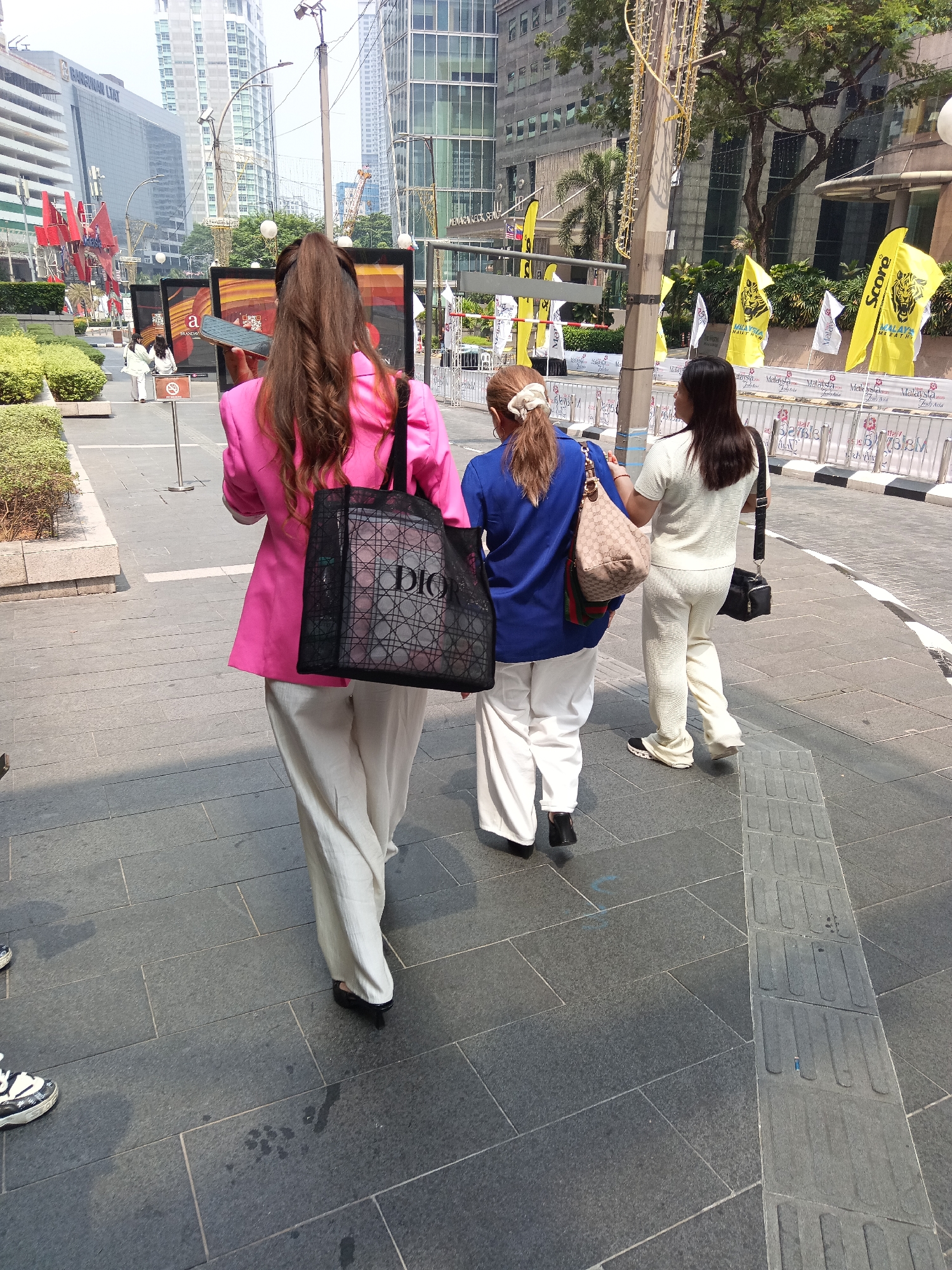 The image captures a street scene in Kuala Lumpur, Malaysia, likely in a modern business district given the high-rise buildings in the background. Three individuals are walking away from the camera on a paved pedestrian walkway.
The person in the foreground is wearing a bright pink blazer over a white top and loose-fitting white pants. They are carrying a large black mesh tote bag with the "DIOR" logo prominently displayed. Their long, brown hair is pulled back into a ponytail.
Behind them, a person in a blue top and white pants is walking, carrying a beige shoulder bag. To their right, a third person is wearing a white textured top and matching white pants, with a black crossbody bag slung over their shoulder.
The environment features urban elements: a paved sidewalk with tactile paving, modern buildings with glass facades, some greenery including bushes and trees, and flags or banners mounted on poles in the background. The lighting suggests it is daytime, with shadows cast on the ground.