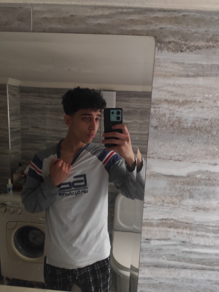 A young male individual, appearing to be in his late teens or early twenties with dark curly hair and dark eyes, is captured in a mirror selfie within a bathroom setting. He wears a long-sleeved raglan shirt with a white torso, gray sleeves, and blue and red stripes on the shoulders, paired with black and white plaid pants. The subject holds a dark-colored smartphone with a distinctive camera module in his right hand, facing the mirror to take the photograph. His left hand grasps the collar of his shirt, pulling it slightly open, while his lips are subtly puckered. The mirror reflects a portion of the bathroom behind the camera, revealing light-colored, horizontally striped wall tiles, a white front-loading washing machine with containers on top to the left, and a white ceramic toilet with a closed lid to the right. The location is an indoor space within As-Salihiya Municipality, Syria.