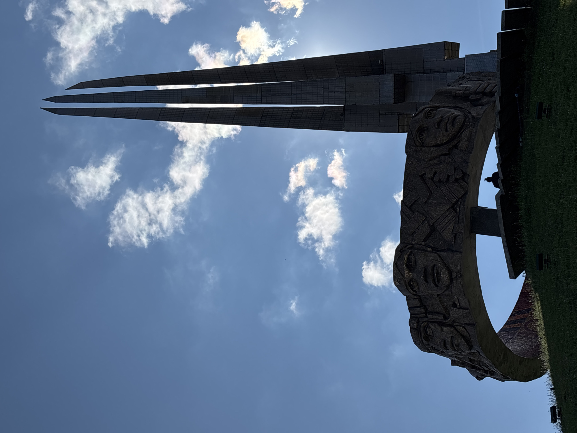 The image shows a war memorial in the city of Ozyaritska-Slabadski rural council, Belarus. The memorial features a large, circular sculpture with carved faces of soldiers, representing different branches of service. Behind this, three tall, slender obelisks rise towards a bright blue sky filled with scattered white clouds. The sun is partially visible, creating a lens flare effect between the obelisks. A lone figure is silhouetted against the sky, seated in the foreground on a grassy slope. The memorial is constructed from concrete and stone, with textured surfaces. The overall scene is one of solemn remembrance, set against a backdrop of a clear, sunny day.