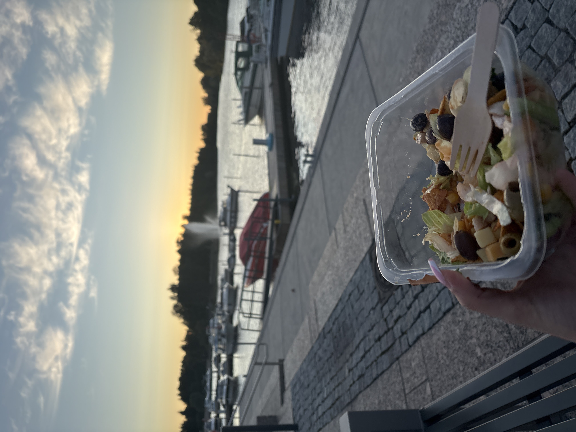 A person is holding a clear plastic container of salad with a wooden fork in it. The container is in the foreground, and the salad appears to contain lettuce, olives, cheese cubes, and possibly some croutons or crackers. The person's hand, with painted nails, is visible holding the container. In the background, there is a body of water, likely the sea or a large lake, with numerous boats docked. A fountain is visible in the water in the middle ground. The sky is overcast with wispy clouds, and the sun is setting, casting a warm orange glow on the horizon. The scene is set at a harbor or marina. Given the context of Pori, Finland, this location is likely Yyteri beach and its marina, known for its maritime activities and proximity to the sea. The overall atmosphere is serene and peaceful, with the warm sunset light creating a tranquil ambiance.