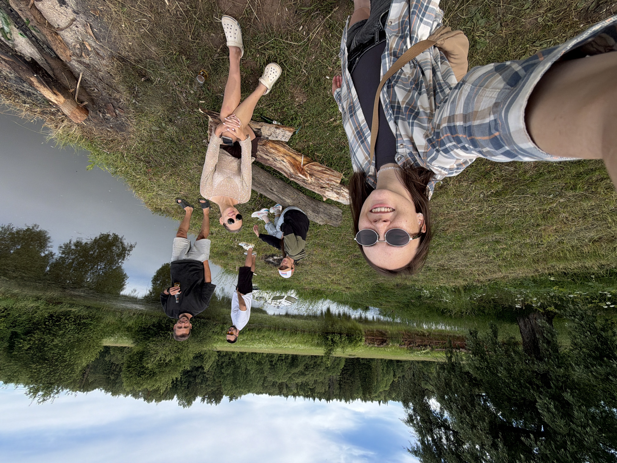 In Garevaya, Russia, five individuals are gathered near a body of water under a cloudy sky. In the foreground, a woman with sunglasses and a plaid shirt is taking a selfie. To her right, another woman is seated on logs, wearing a light-colored top and shorts. Behind them, three men are standing. One man in a white t-shirt and shorts is looking away, while another in a black t-shirt and shorts holds a bottle. A third man, partially visible, is also present. The environment is natural, with a grassy bank leading to the water, surrounded by trees and foliage. A small white object, possibly a boat, is visible in the water. The scene appears to be a casual outdoor gathering.