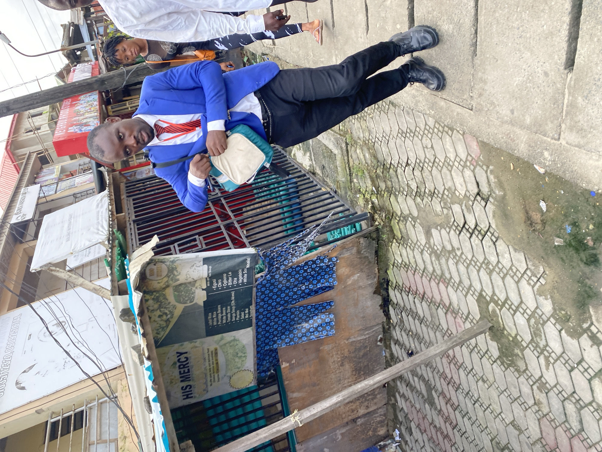 A man in a blue suit, white shirt, and red tie is walking on a sidewalk next to a street market stall. He is carrying a light-colored bag over his left shoulder. He is wearing black pants and black shoes. To his right, a woman with dark curly hair and a black top with a pattern is walking in the same direction. Further to the right, a person in a white shirt is partially visible. The market stall has a corrugated metal roof and a wooden front with a sign that reads "HIS MERCY." Blue patterned fabric is hanging on the stall. The ground is paved with interlocking stones, and there are puddles of water and some debris. Buildings and more market stalls are visible in the background.