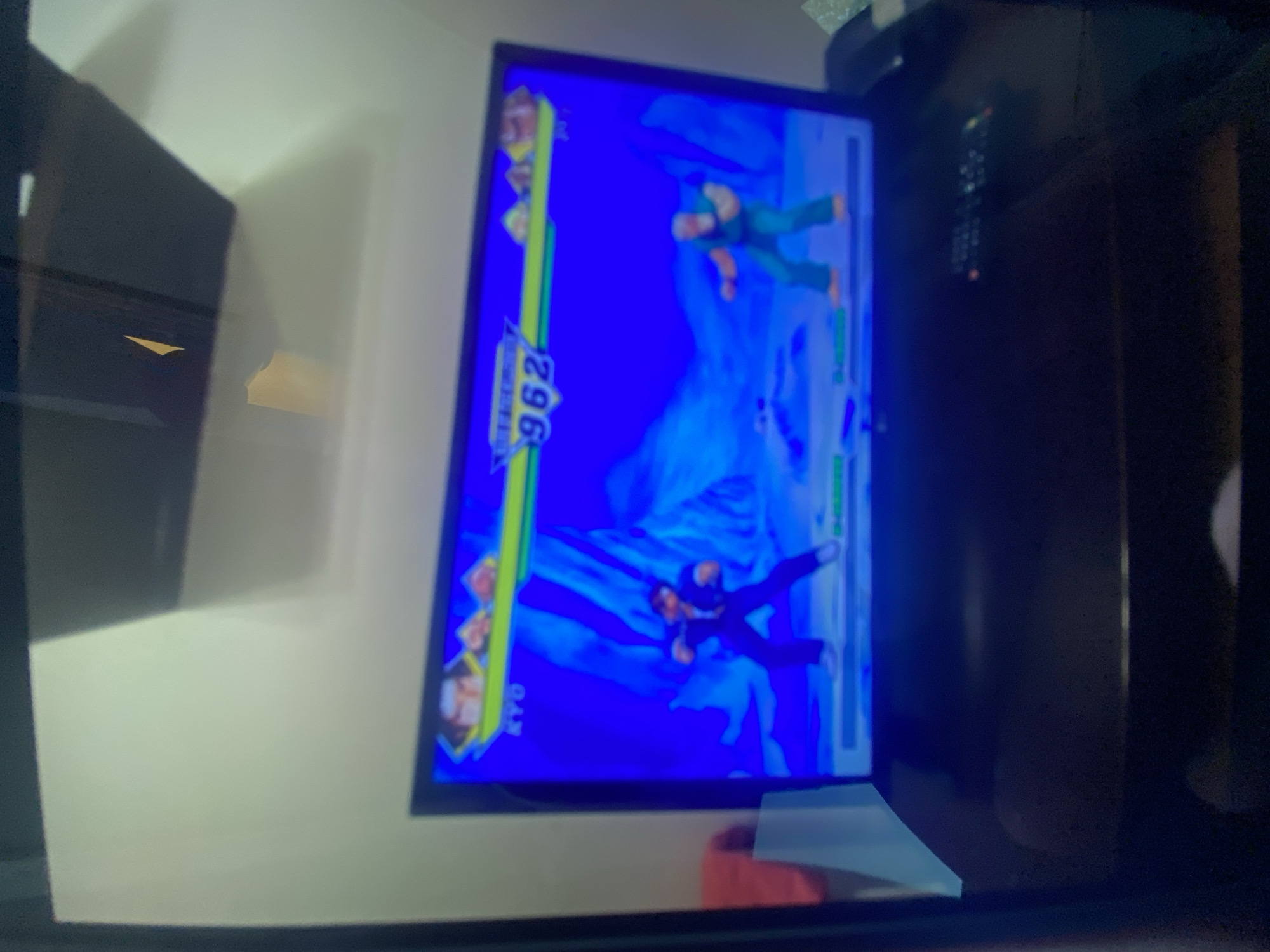 A television displays a fighting video game featuring two characters in mid-action. The character on the top right is wearing green clothing, while the character on the bottom is in black clothing. Numbers and meters are visible on the display. The television is situated on a stand in a room with a white wall on the left. The environment is dimly lit. There is no evidence suggesting the location is Plovdiv, Bulgaria.