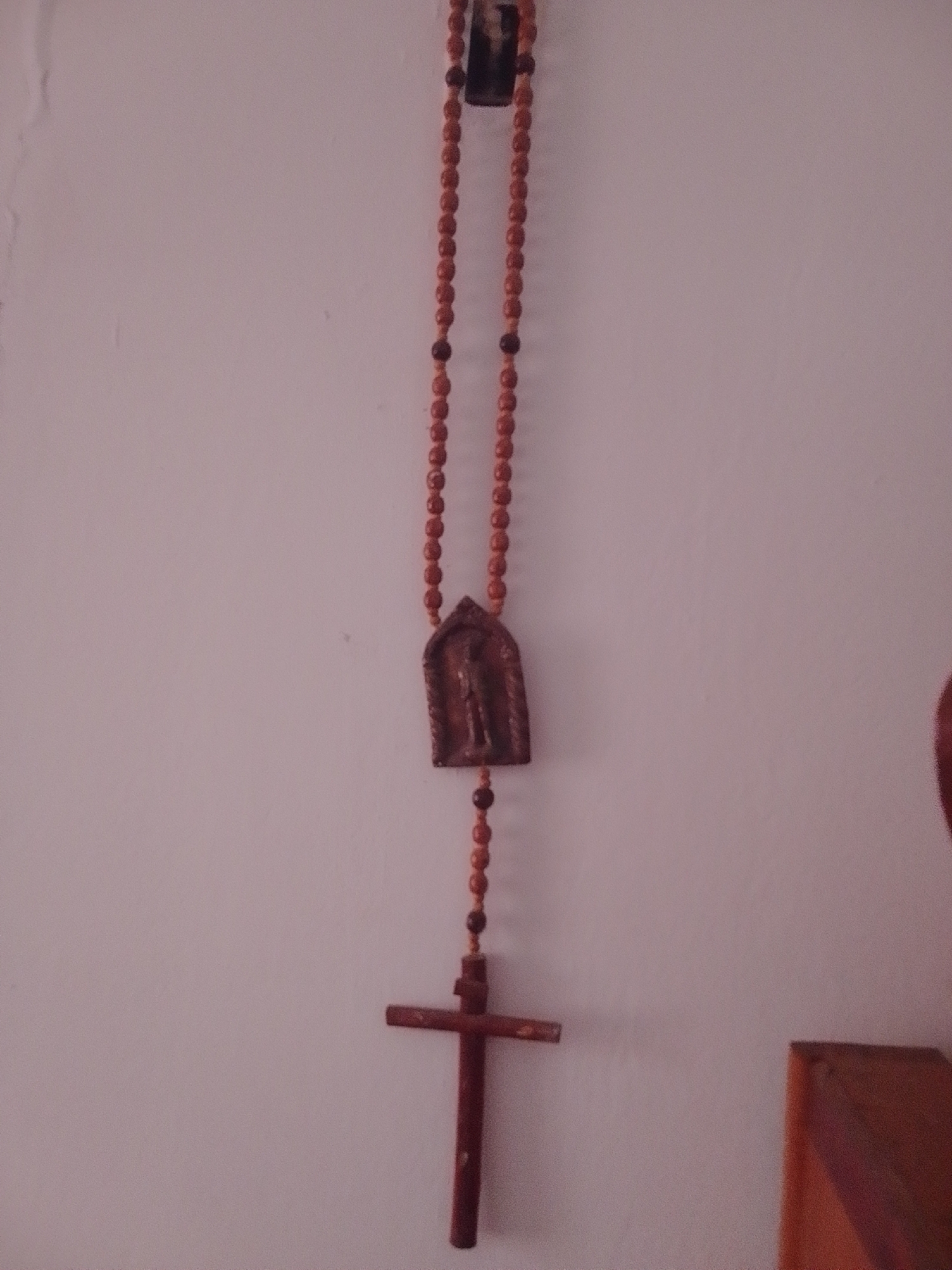 This media file shows a close-up of a religious object, specifically a rosary, hanging against a plain, light-colored wall, likely indoors. The rosary is made of small, reddish-brown wooden beads strung together. At the top, a small, darker wooden crucifix-like charm with an image of a figure is visible. Further down the strand of beads, there is a larger, darker wooden cross. The lighting suggests it is daytime, as the light source is casting a soft illumination on the wall and the object. The overall scene is simple and focused on the religious item, with no other objects or people clearly visible in the frame. The setting appears to be a domestic or devotional space, given the nature of the object. There is no visible text in the image.