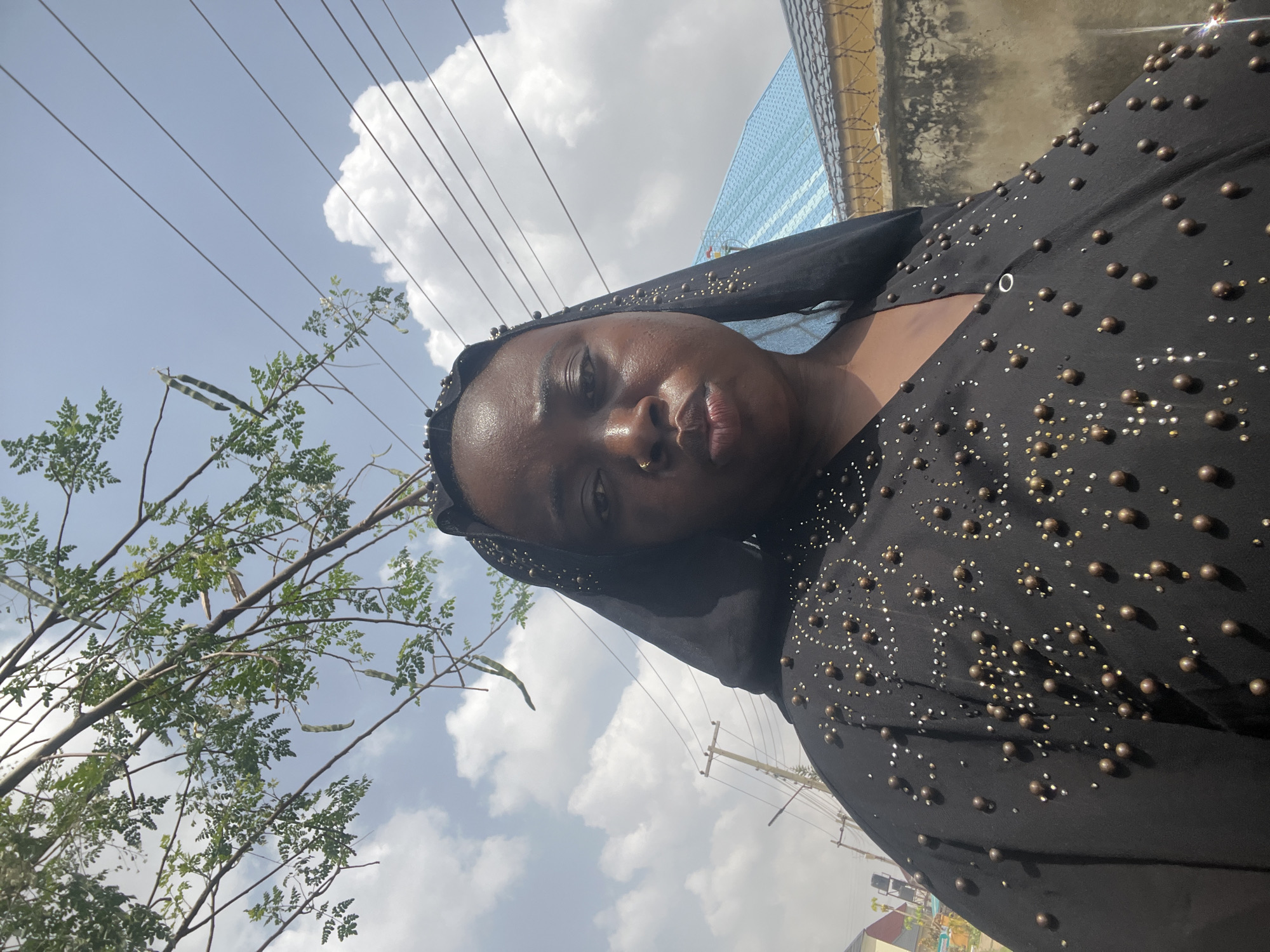 A medium-shot, tilted, outdoor photograph depicts a Black woman wearing a black hijab and black top with beaded detailing. She has dark skin, brown eyes, a nose ring, and full lips. She is looking directly at the camera with a slightly pouting expression. The background includes a blue sky with scattered clouds and some leafy tree branches on the left. Electrical wires are seen extending across the upper part of the image. Buildings and a fence are visible in the background. The photograph appears to have been taken in Abuja, Nigeria, as indicated by the urban setting, electrical poles, and architectural details.