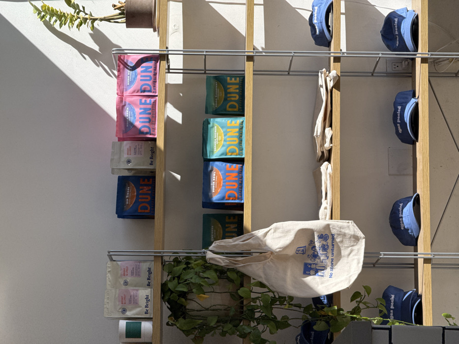 This image depicts an interior retail display within a coffee shop or specialty store in Los Angeles, California, United States. A multi-tiered shelving unit, constructed with light wood shelves and thin metal uprights, is positioned against a white wall.
The top shelf on the left features a cylindrical green container next to two white pouches labeled "Be Bright," identified as "Bright & Lively" coffee. To their right, two dark blue pouches are labeled "DUNE Coffee Roasters," specifically "Zip Fingers." Further right, two more white "Be Bright" pouches precede two pink pouches, also "DUNE Coffee Roasters," identified as "Diego Horta Colombia." A potted plant with a single upright stem and green leaves is on the far right.
The middle shelf displays a trailing green plant (likely a Pothos) in a pot on the far left. Hanging from the metal support to its right is a beige canvas tote bag with a blue graphic depicting figures and text "No stone unturned." To the right of the tote bag, four "DUNE Coffee Roasters" pouches are visible: a dark green "Tejeda" pouch, an orange "Punch Bowl" pouch, a teal "Hispania Guzman Honduras" pouch, and another dark green "Tejeda" pouch. On the far right of this shelf, several light-colored folded fabric items are stacked.
The bottom shelf on the left shows two blue baseball caps, with one partly obscured, each embroidered with "eyes peeled" in white text. To their right, another three identical "eyes peeled" blue baseball caps are displayed. An electrical outlet is visible on the white wall section behind the rightmost caps.
Natural light casts sharp diagonal shadows across the white wall and shelving, indicating a sunny environment. No individuals are present in the frame. The overall scene suggests a commercial establishment selling coffee beans and branded merchandise. The specific "DUNE Coffee Roasters" branding indicates a location associated with this company, which operates in Los Angeles.