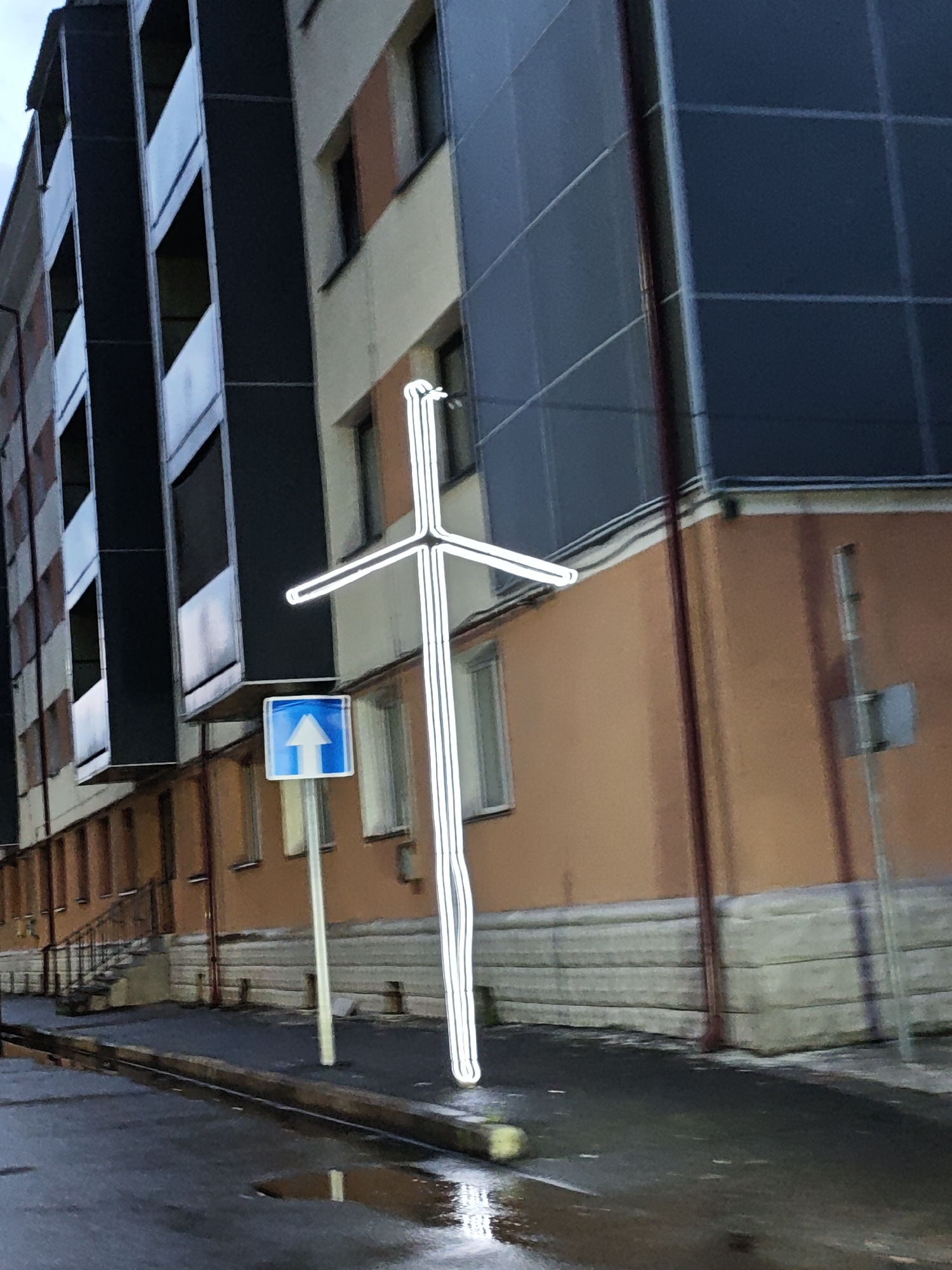 In Tqibuli, Georgia, an illuminated cross stands in front of a building. The cross is a vertical structure with a horizontal beam near the top, outlined in white, suggesting it is made of lights or reflective material. To the left of the cross, a blue and white directional sign with an upward-pointing arrow is mounted on a pole. The building behind them features a dark, patterned facade on the upper floors, contrasting with a light-colored section and an orange-colored lower section. The ground in the foreground is wet, with puddles reflecting the surrounding environment, indicating recent rain or damp conditions. A narrow street or sidewalk is visible, with a curb separating it from the road. The overall scene appears to be an outdoor urban setting, possibly at dusk or during overcast weather, given the lighting.