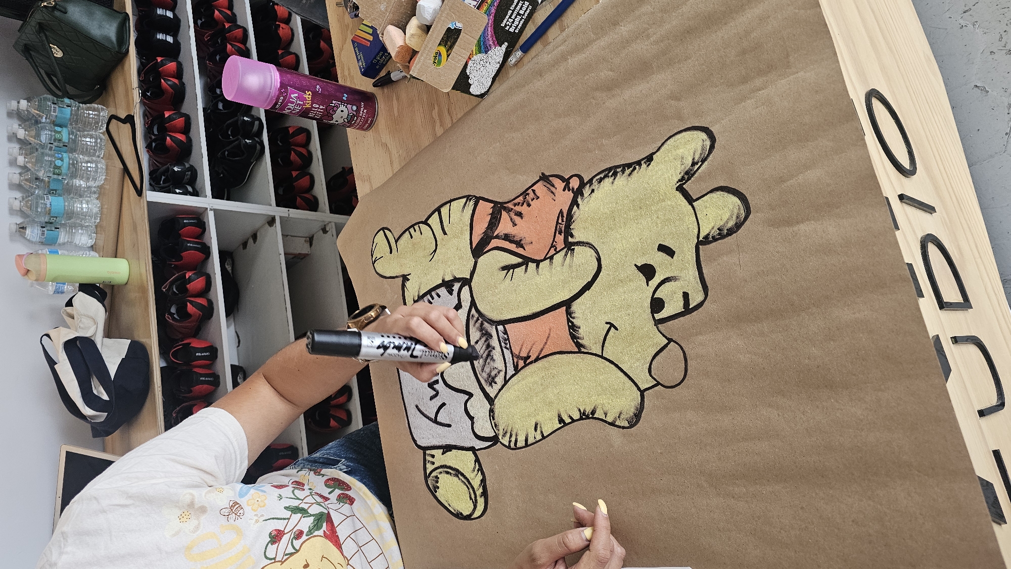 A person is drawing Winnie the Pooh on brown paper using a black marker. The scene appears to be indoors, possibly a workshop or studio, with various items such as water bottles, shoes, a purse, art supplies, and a wooden frame with black letters on it visible in the background. The person is wearing a t-shirt with a Winnie the Pooh graphic and jeans. The location, based on the OCR text, is possibly Puebla, Mexico, although the exact location cannot be pinpointed.