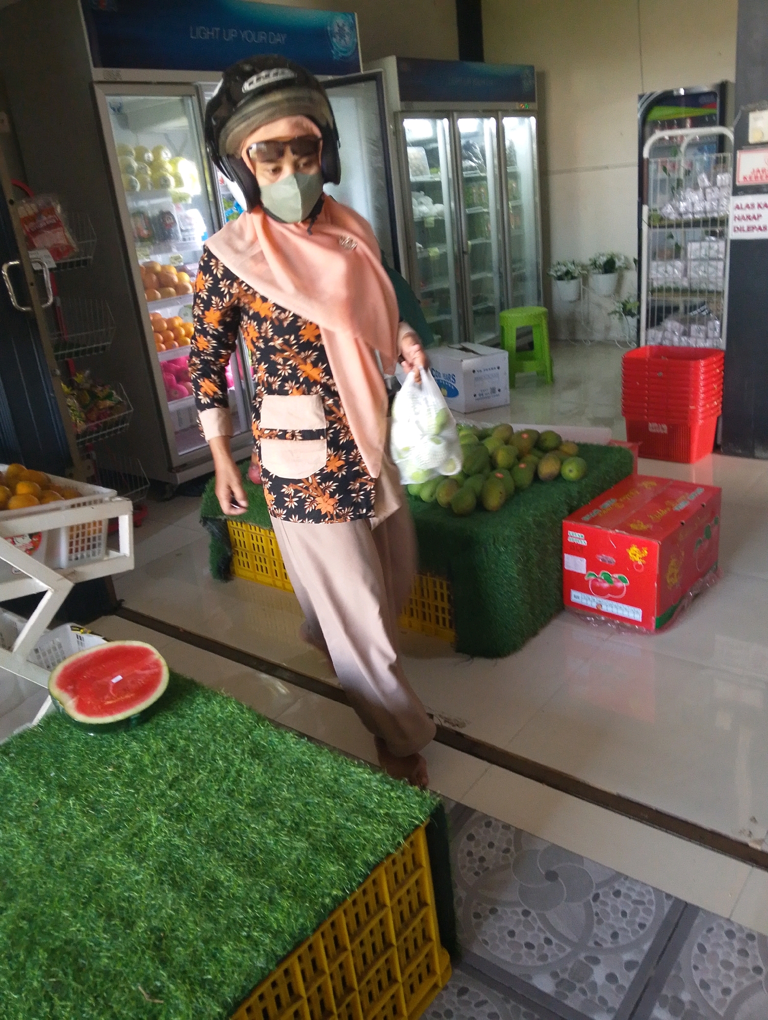 A person wearing a helmet, sunglasses, and a face mask is standing in what appears to be a market or grocery store in Bangil, Indonesia. The person is holding a plastic bag filled with green fruits, likely mangoes, and is wearing a patterned shirt and light-colored pants. In the foreground, a slice of watermelon is displayed on a green artificial grass surface, positioned on a yellow plastic crate. Behind the person, a refrigerator unit filled with fruits is visible, along with shelves displaying various items. Several red plastic baskets are stacked to the right of the scene, and a red cardboard box with an image of apples is placed on the floor. The environment suggests a retail setting with fresh produce and goods on display.
