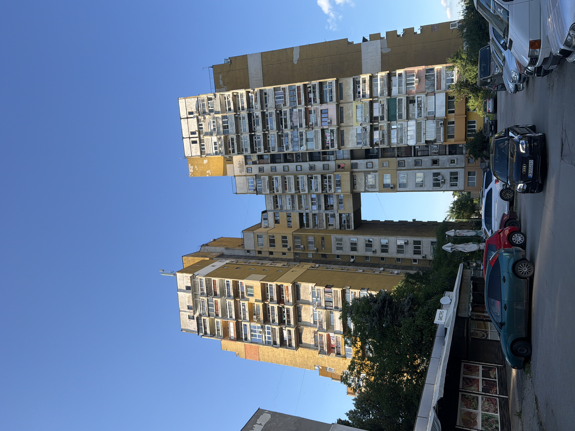 The image shows a distinctive residential building in Sofia, Bulgaria, characterized by its unique architecture featuring a large archway connecting two towers. The building is predominantly yellow and gray with numerous balconies and windows. The sky is clear blue with a few white clouds. In the foreground, there is a parking lot with several cars, including a black Audi, a red car, and a green hatchback. Adjacent to the parking lot is a small building with a canopy, appearing to be a food establishment with pictures of dishes displayed on its windows. Trees and foliage are visible around the building and in the background. The scene is captured from a low angle, emphasizing the height and scale of the residential complex.