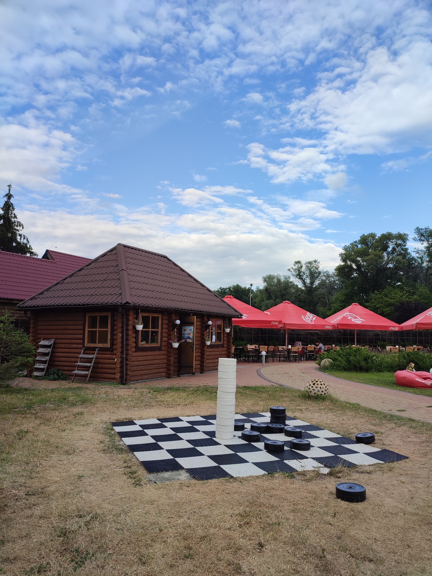 The image depicts an outdoor recreational area with a wooden building, a checkerboard game, and an outdoor dining area. The wooden building, possibly a cafe or restaurant, has a brown, shingled roof and is constructed from logs. Two ladders lean against its side. In front of the building is a large checkerboard laid out on the ground, with several black checker pieces scattered on it. To the right of the checkerboard, there's a red beanbag chair with the Coca-Cola logo. Beyond that, a paved path leads to an outdoor seating area covered by large red umbrellas, also bearing the Coca-Cola logo. Several tables and chairs are set up under the umbrellas, and some patrons are seated. The background is filled with lush green trees. The sky is blue with scattered white clouds. The location is identified as Shcherbani, Ukraine.