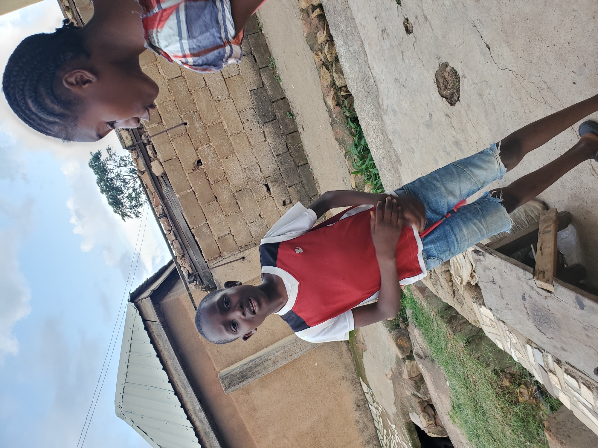 A young boy and an older girl stand in an outdoor urban setting in Jos, Nigeria. The boy, facing the camera, wears a white t-shirt with a red and black sleeveless vest and denim shorts. His hands are clasped in front of him. The girl, to the right and partially out of frame, has braided hair and wears a plaid shirt. Behind them, a weathered brick wall separates the area from buildings with corrugated metal roofs. The ground is a mix of concrete and dirt, with patches of grass and some rocky debris. A wooden structure is visible in the lower left foreground. The sky is partly cloudy.