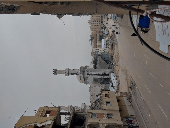 The image captures a wide-angle view of a heavily damaged urban street scene in Khan Yunis, Palestinian Territory, under an overcast sky.
In the center, a large mosque with a prominent central dome and two minarets is severely structurally compromised; its main prayer hall appears largely collapsed, surrounded by significant piles of rubble. One minaret stands mostly intact, while the other's upper section is missing.
To the left, a multi-story building is extensively damaged, with multiple floors exposed due to missing walls and shattered windows. Visible rebar protrudes from the fractured concrete. Another mosque-like structure or part of the main mosque's minaret is visible behind this building. The ground level displays debris and faded graffiti.
To the right, another multi-story building shows comparable extensive damage, with its facade partially removed, revealing interior spaces. In the immediate foreground on the right, thick black cables and wires hang, partially obscuring a makeshift blue structure and additional debris.
A wide asphalt road with clear dashed white lane markings runs through the middle ground, appearing largely clear of large debris. However, the verges and areas surrounding the damaged structures are covered in extensive rubble and dirt.
Approximately 10-15 individuals are dispersed along the road in the mid-distance, walking in various directions, primarily moving away from the viewer. One person in dark clothing walks on the right side of the road closer to the foreground. In the background, further down the street, a cluster of multi-story buildings, some appearing less damaged, are visible, along with a large white tent-like structure.