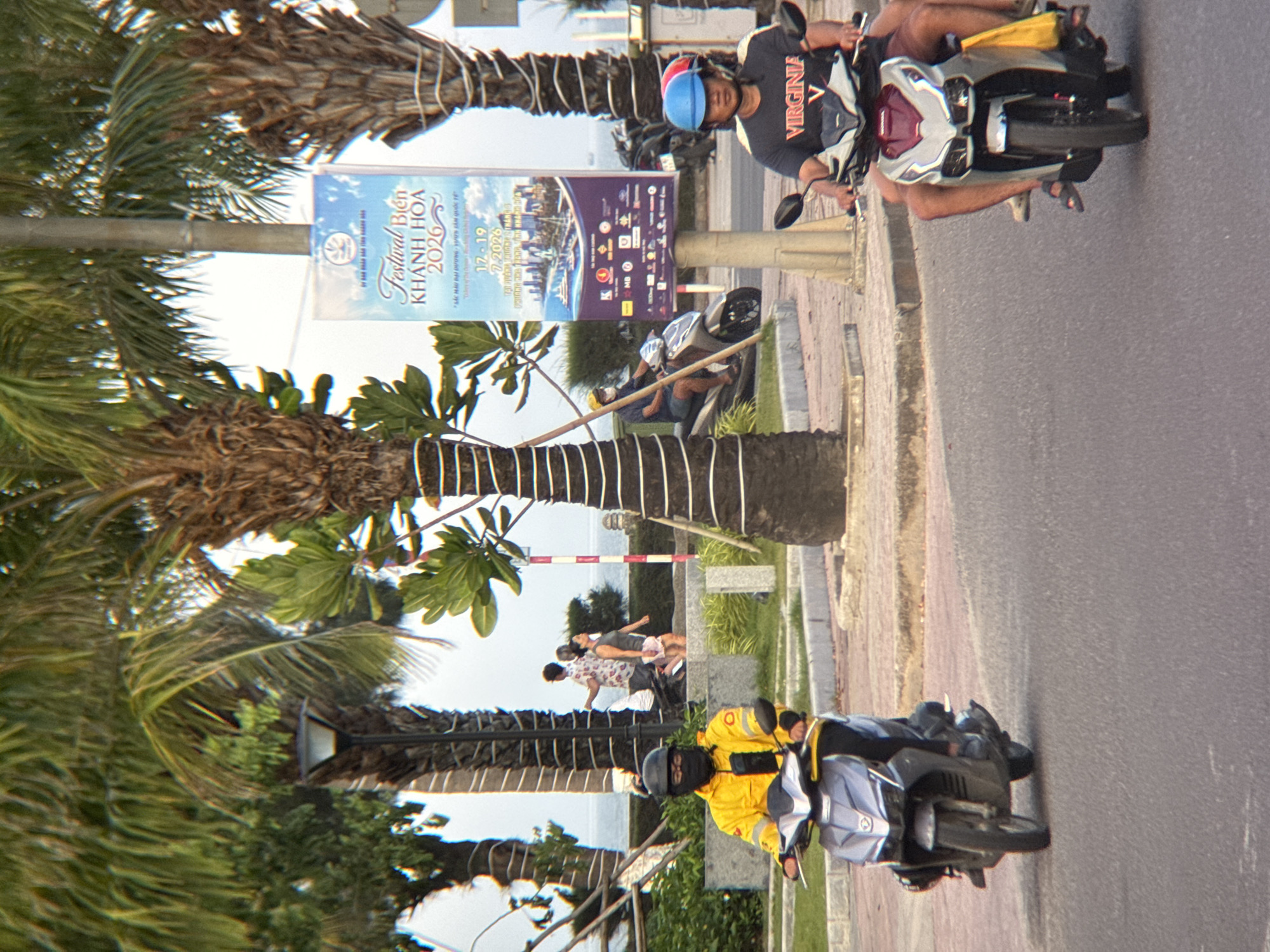 In Nha Trang, Vietnam, two individuals are riding motorcycles along a street lined with palm trees. The person in the foreground on the right is wearing a black t-shirt with "VIRGINIA" printed on it and a blue helmet. They are riding a silver and red motorcycle. To the left, another person is riding a silver motorcycle, wearing a yellow jacket, black pants, and a black helmet and face covering. In the background, a poster advertises "Festival Bien Khanh Hoa 2026". Further back, several people are walking on a sidewalk near the ocean, with palm trees and a lamppost visible. The scene appears to be set in a coastal urban environment.