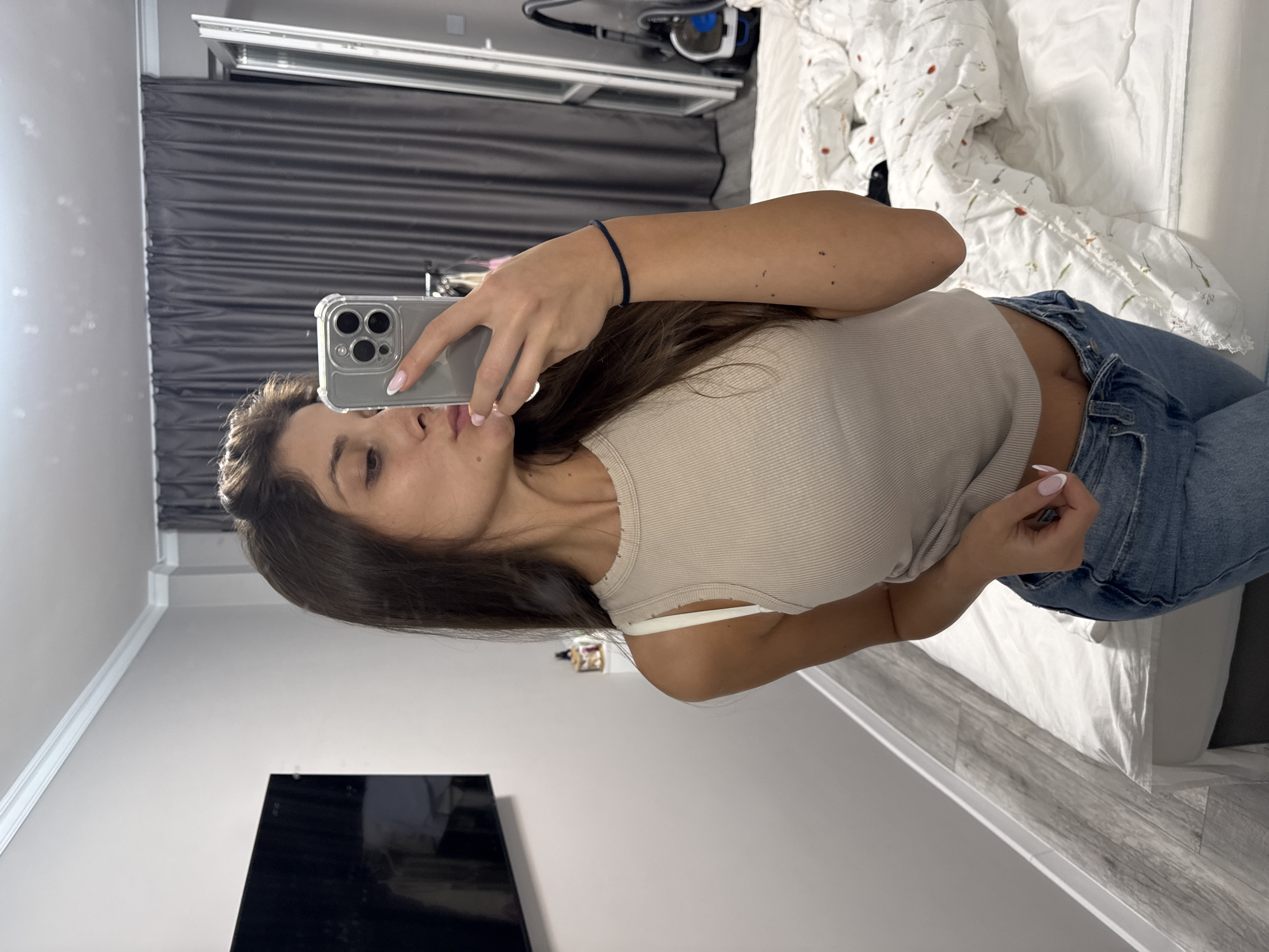 A woman is taking a selfie in a bedroom. She is holding a phone with a clear case and two rear cameras. She has long brown hair and is wearing a beige tank top and jeans. The bedroom has a bed with a white comforter with floral patterns. There is a gray curtain in the background, as well as a television. The location cannot be determined to be in Burgas, Bulgaria from the given image.