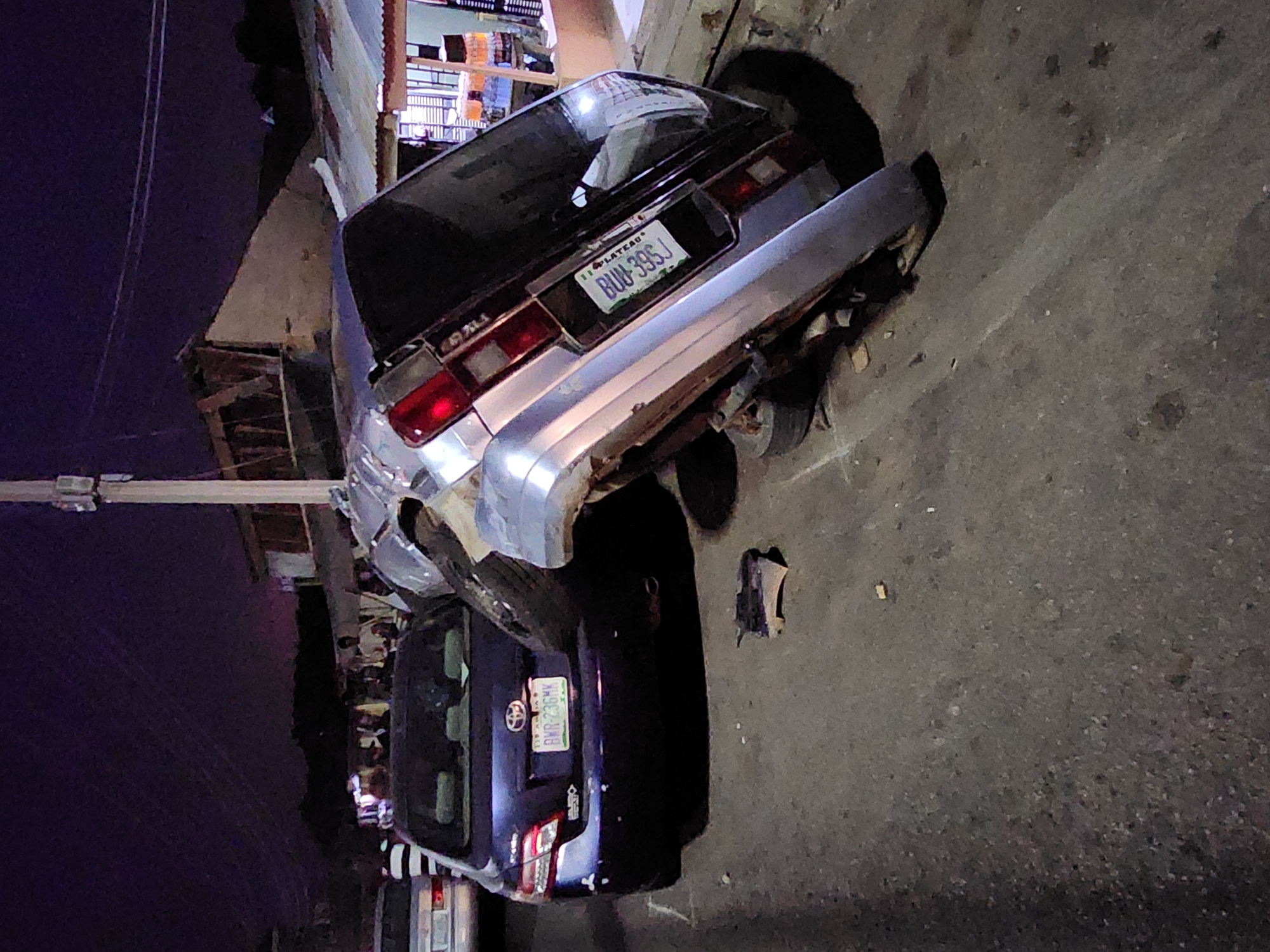The image shows two cars involved in an accident on a street in Unknown, Unknown. One car, a silver or light-colored sedan with the license plate "BUW-39SJ PLATEAU", is angled upwards. A dark-colored car is directly below it, facing the opposite direction. The license plate of the second car is visible, reading "BWR-236MK". A crowd of people is gathered in the background, partially obscured by shadows. A pole is visible on the left side of the frame. The ground is paved.