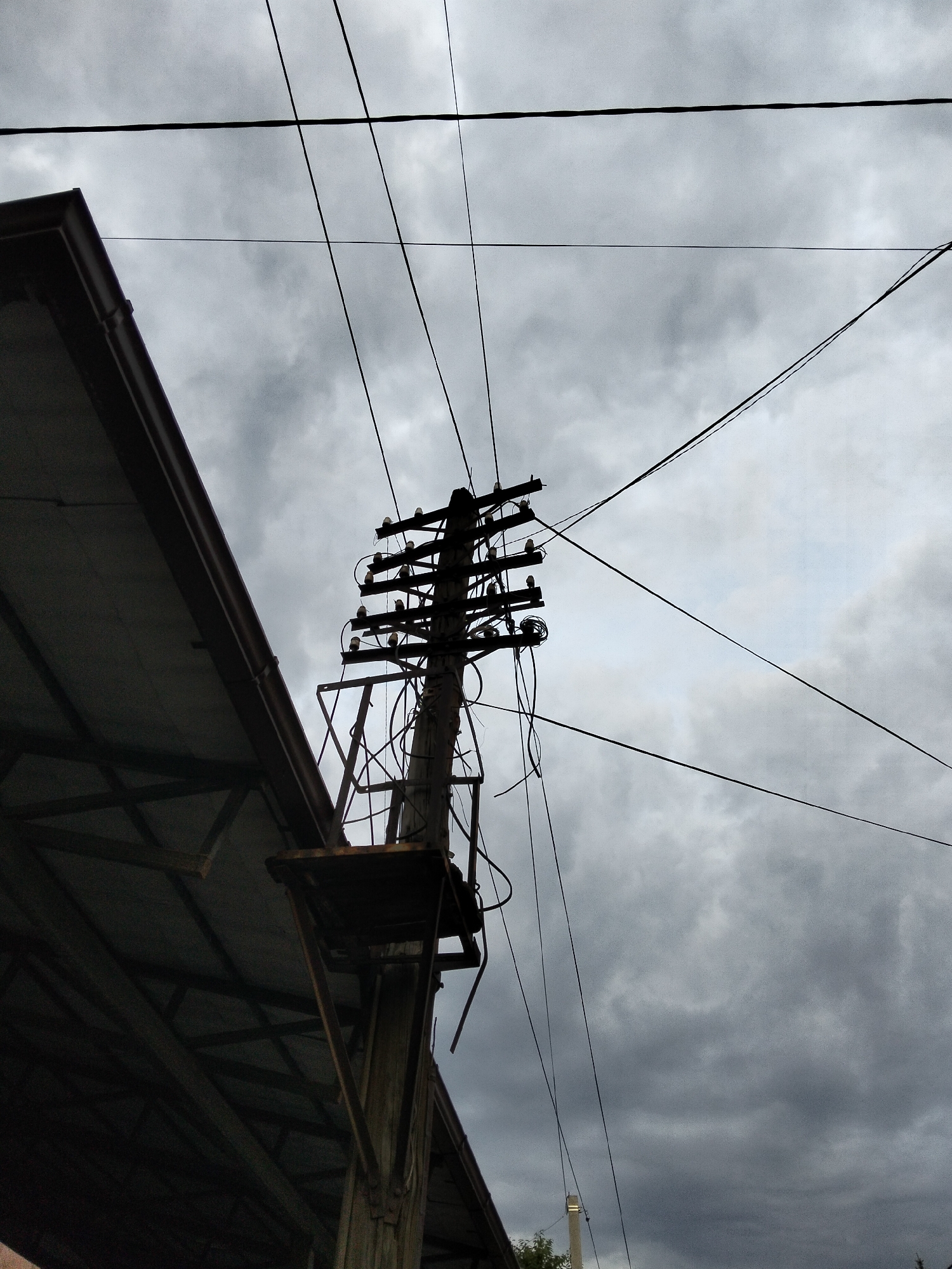 This media file shows a low-angle shot of a utility pole against a cloudy sky. The utility pole, made of wood, is adorned with multiple crossbars holding ceramic insulators and electrical wires. Some wires are taut, while others hang loosely, suggesting a somewhat disorganized setup. A metal platform is attached to the side of the pole. To the left of the pole, the dark, angular roofline of a building with visible structural beams is prominent. The sky is overcast with heavy, gray clouds, indicating a gloomy or possibly rainy day. The overall scene evokes a sense of an urban or semi-urban environment, possibly in Almaty, Kazakhstan, given the location context. The time of day is indeterminate due to the cloud cover, but the lighting suggests it is daytime. There is no discernible text in the image. The scene is devoid of people, and the focus is on the utilitarian infrastructure and the atmospheric sky.