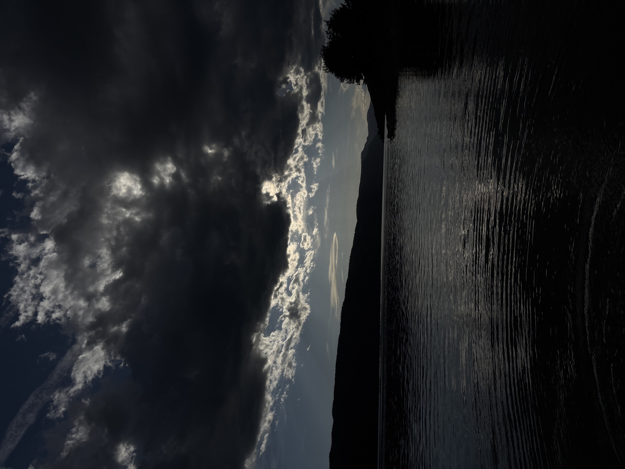 The image depicts a body of water, possibly a lake, under a sky with dramatic cloud cover. Heavy, dark clouds dominate the left side of the image, while the right side features more fragmented clouds that allow glimpses of sunlight. A dark, silhouetted landmass runs along the center of the frame, separating the sky from the water. The water surface is subtly textured, suggesting gentle ripples or waves. The extreme right of the image is almost entirely dark. The location in the city of Unknown, Unknown is not determinable from this single image.