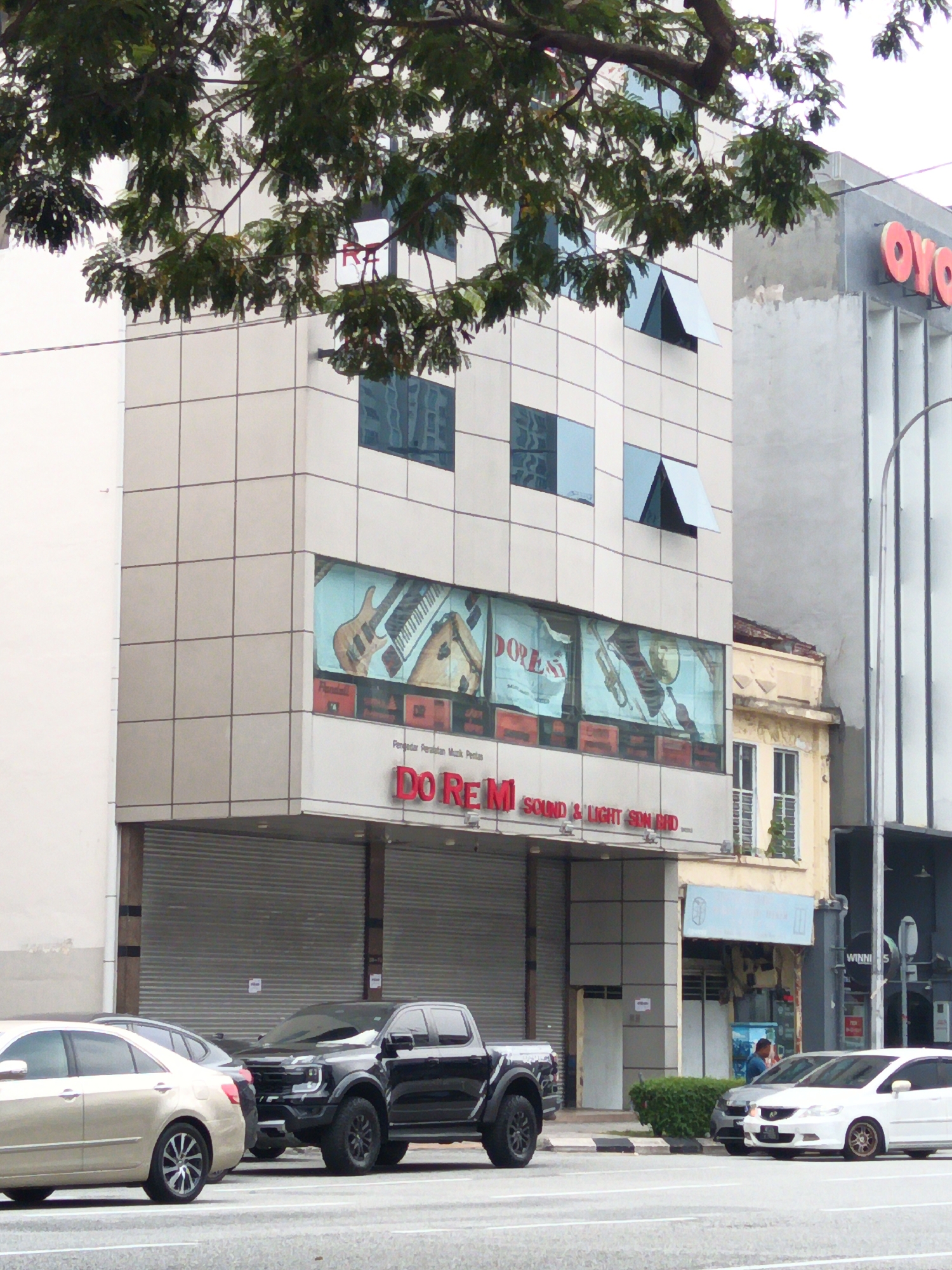 This image captures a street view in Kuala Lumpur, Malaysia, focusing on a multi-story building with a distinctive facade. The building on the left is clad in light-colored square tiles and features several windows, some of which are open and angled outwards. A large mural depicting musical instruments, including guitars and keyboards, adorns a prominent section of the building's lower floors, with the text "Do Re M SOUND & LIGHT SDN BHD" visible below it. To the right of this building, another structure with a yellow facade and windows is visible, and further to the right, part of a modern building with vertical elements and a large "WINNI" sign is present. In the foreground, several vehicles are parked on the street or on a sidewalk. A light-colored sedan is on the left, followed by a black pickup truck with aggressive styling. A white hatchback is parked further to the right. The street is paved, and a pedestrian walkway with some greenery is visible. Trees with dense foliage partially obscure the upper portions of the buildings. The overall scene suggests a commercial area.