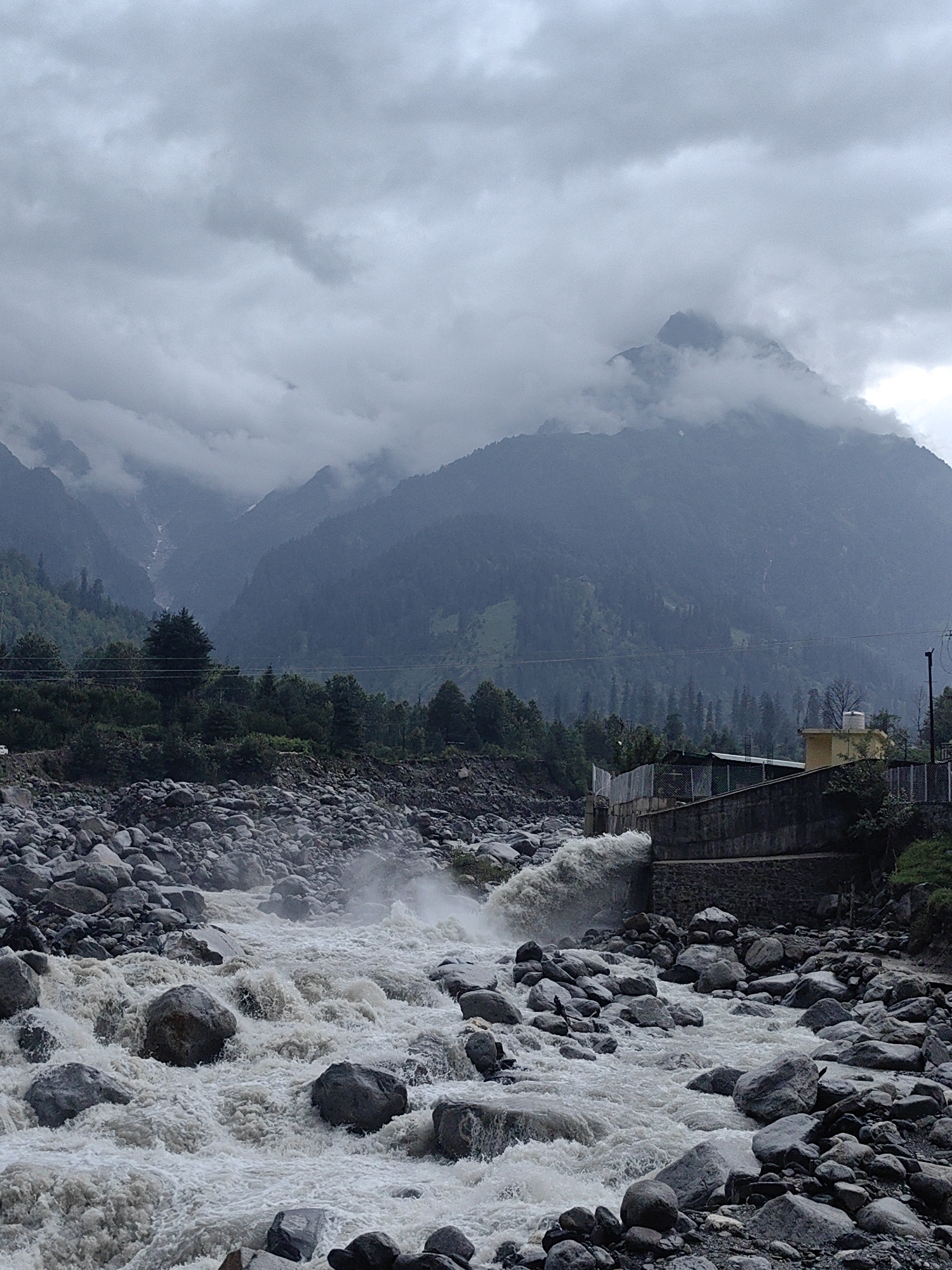 A turbulent river flows through a rocky gorge, with a concrete structure and a small yellow building visible on the right bank. In the background, misty mountains covered in trees rise into a cloudy sky. The scene appears to be in the mountainous region of Kulang, India, characterized by its natural landscape and infrastructure.