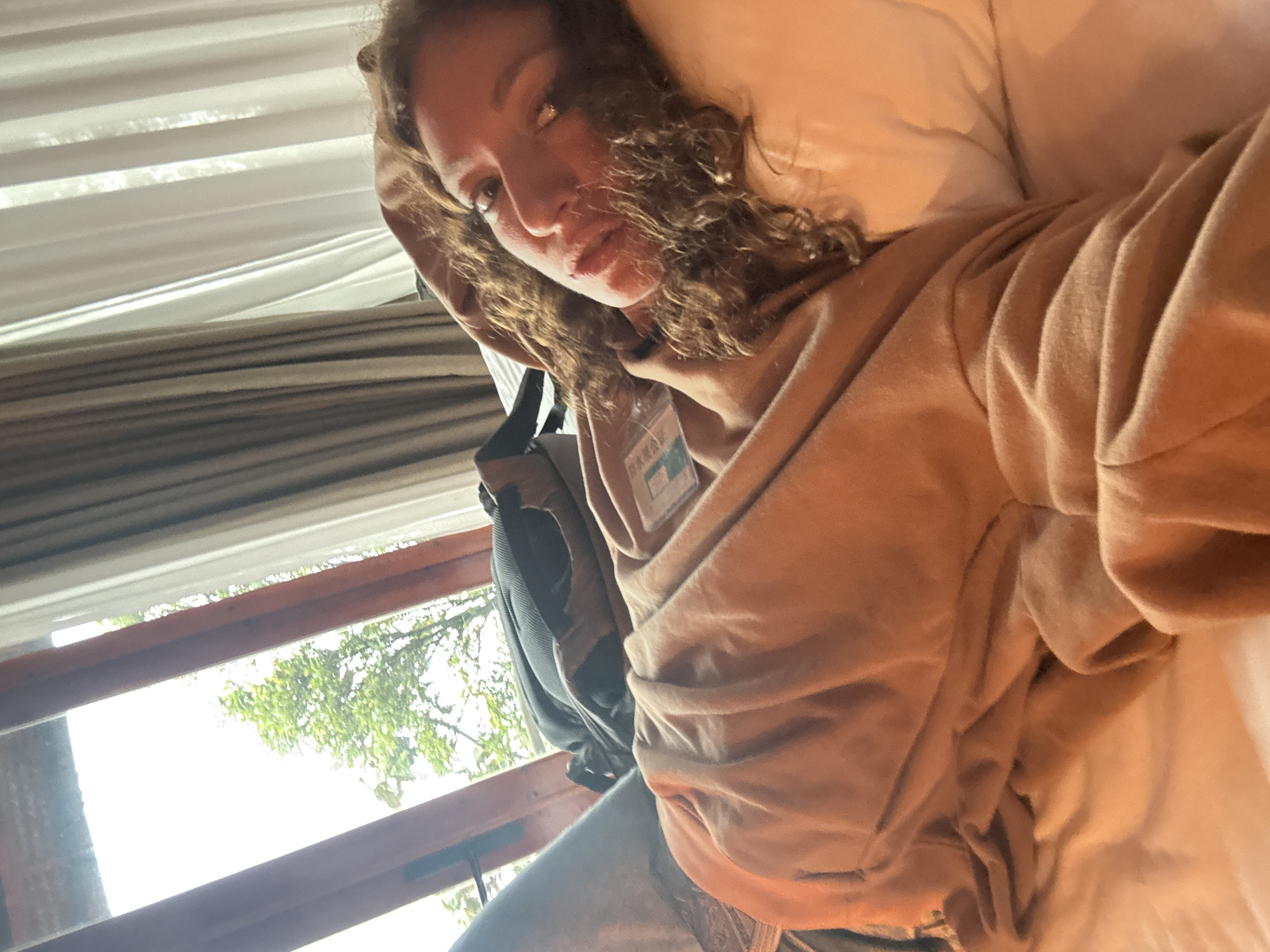 A woman with curly brown hair is lying in a bed. She is wearing a brown hoodie and has a lanyard around her neck. A backpack is partially visible next to her. A window is in the lower left of the frame, showing green foliage outside. The location is likely an indoor space in Binh Lu, Vietnam, as indicated by information on the lanyard.