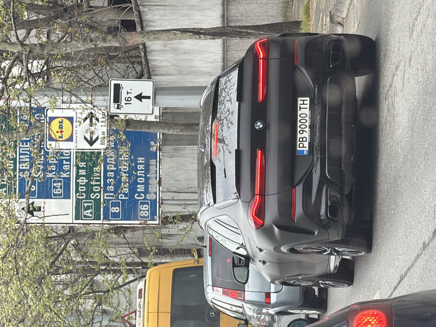 The image depicts a daytime street scene, likely in Bulgaria, with several vehicles halted in traffic. The central focus is a black BMW SUV, possibly an X6, with a matte finish and prominent quad exhaust pipes. Its Bulgarian license plate reads "PB 9000 TH" and includes the country code "BG." Below the license plate, the text "EXCLUSIVE CARS center" is visible. The BMW's red taillights are illuminated.
Ahead of the BMW, a silver or grey SUV is partially visible, and behind it, a yellow van can be seen. In the immediate foreground, to the right, the red taillight of another dark-colored car is present.
To the left of the vehicles, a large blue road sign provides directions to various Bulgarian cities, written in both Cyrillic and Latin scripts. Destinations listed include "А1 София" (A1 Sofia), "8 Пазарджик" (Pazardzhik), "86 Смолян" (Smolyan), and "64 Карлово" (Karlovo). The sign also features the "LIDL" logo and indicates distances of "800 м 1,8 км" (800 m 1.8 km). Above it, a white square sign with a truck icon and "16 т." (16 tons) points upwards, suggesting a weight limit or route for heavy vehicles.
Trees with sparse green leaves, indicative of spring foliage, are visible on the left side of the frame. The lighting appears soft, suggesting an overcast day. All vehicles are stationary, implying they are stopped in a traffic queue.