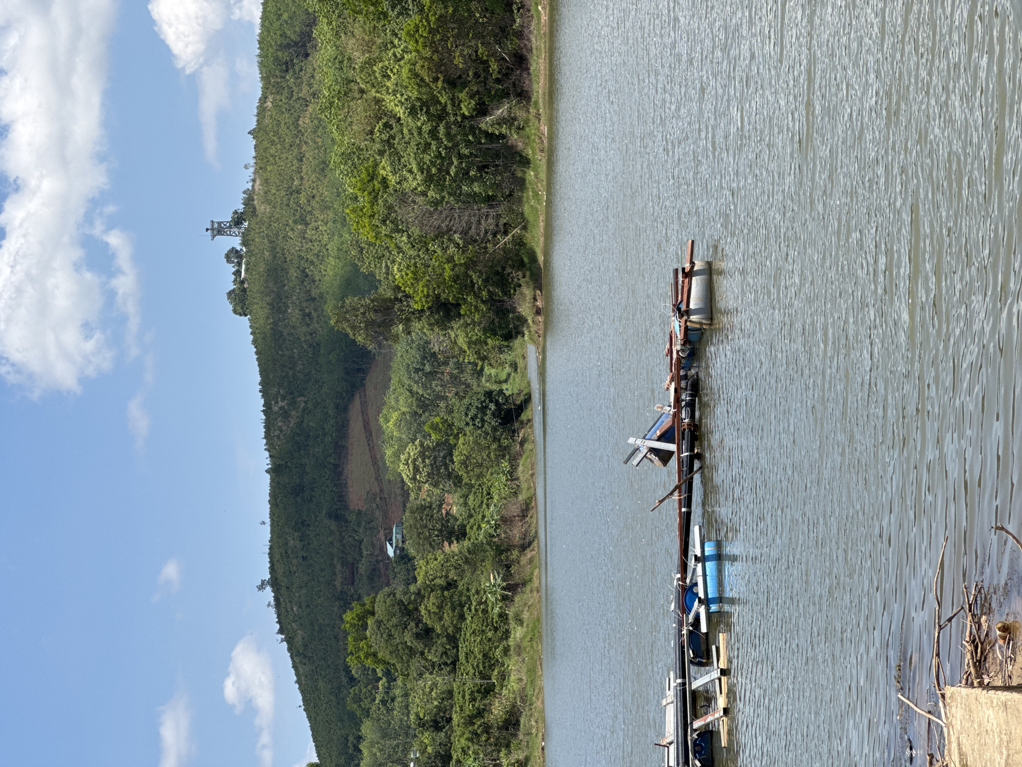 The image captures a waterscape in Bờ Y, Vietnam, featuring a body of water adjacent to a densely wooded hillside. A transmission tower is visible atop the hill. An assemblage of makeshift floating structures rests on the water's surface, constructed from barrels, planks, and pipes. The sky is partially obscured by scattered clouds. The terrain consists of thick vegetation meeting the shoreline.
