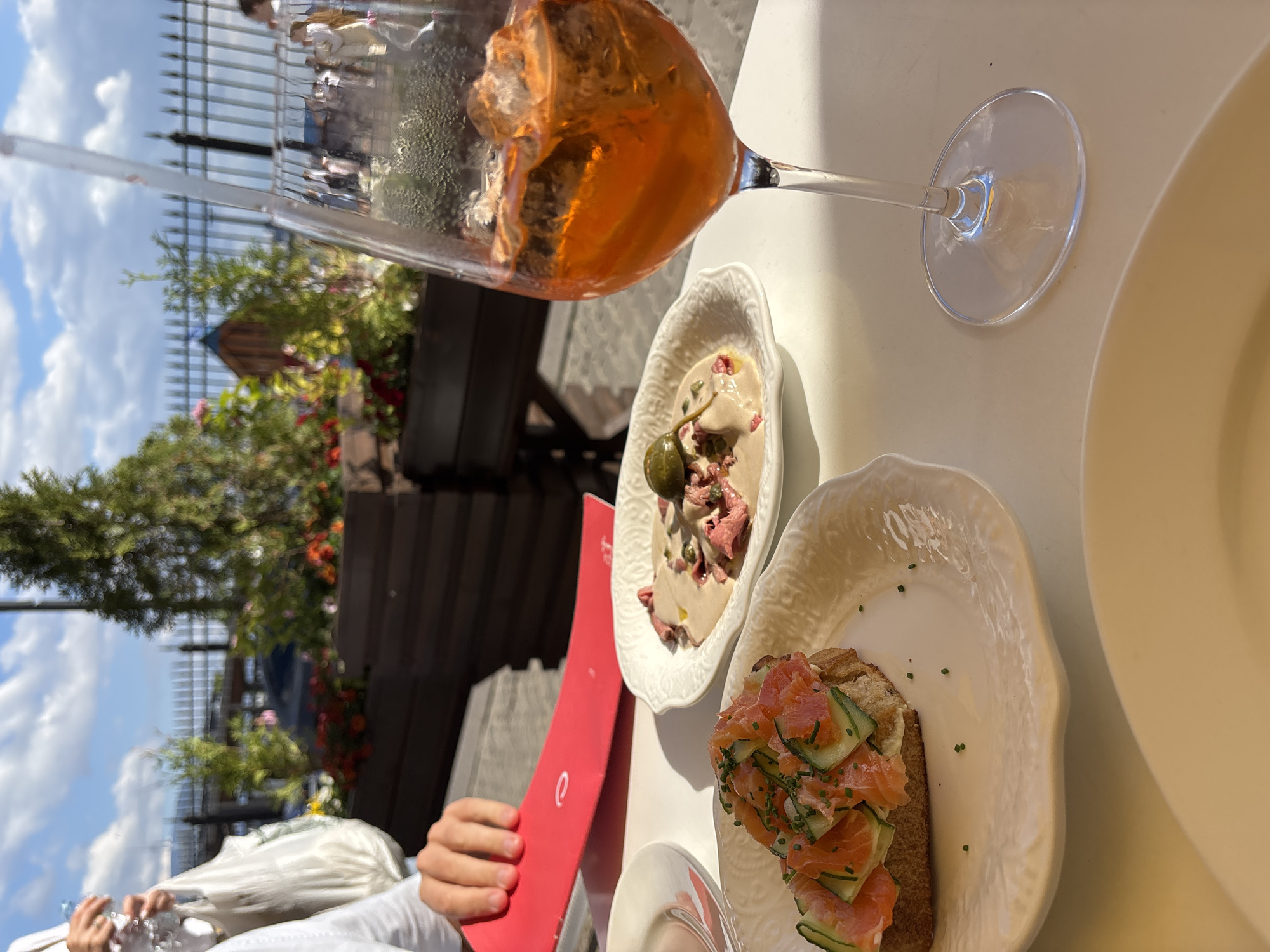The image depicts an outdoor dining scene at a cafe in Saint Petersburg, Russia, with the Peter and Paul Fortress visible in the background. In the foreground, a table is laden with food and drinks. A glass of an orange aperitif, likely Aperol Spritz, sits prominently on the right, filled with ice. To its left, a white oval plate holds a dish of thinly sliced meat with a creamy sauce and a caper. Closer to the viewer is a slice of toasted bread topped with smoked salmon, cucumber slices, and chopped chives, presented on another white embossed plate. A hand, presumably belonging to one of the diners, is visible reaching for a red menu. The setting appears to be a sunny day, with a blue sky dotted with clouds. Lush green plants and flowers in wooden planters line a metal fence in the background, suggesting a well-maintained outdoor seating area.