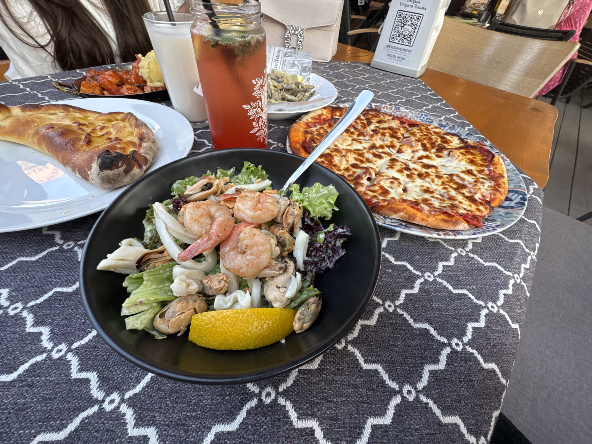 A table is set with a variety of dishes at a restaurant in Primorsko, Bulgaria. In the foreground, a black bowl contains a seafood salad with shrimp, mussels, and calamari, garnished with a lemon wedge. To the right, a pizza with melted cheese and pepperoni is served on a decorative plate. A piece of khachapuri, a Georgian cheese-filled bread, sits on a white plate to the left. Behind the food, two drinks are visible: a tall glass with a red beverage and mint, and another glass with a light-colored drink. A small plate with fried fish and lemon is also present. A QR code menu stands on the table, indicating a digital ordering system. The table is covered with a dark grey tablecloth featuring a white geometric pattern. Two people's torsos are partially visible in the background, suggesting they are dining at the restaurant.