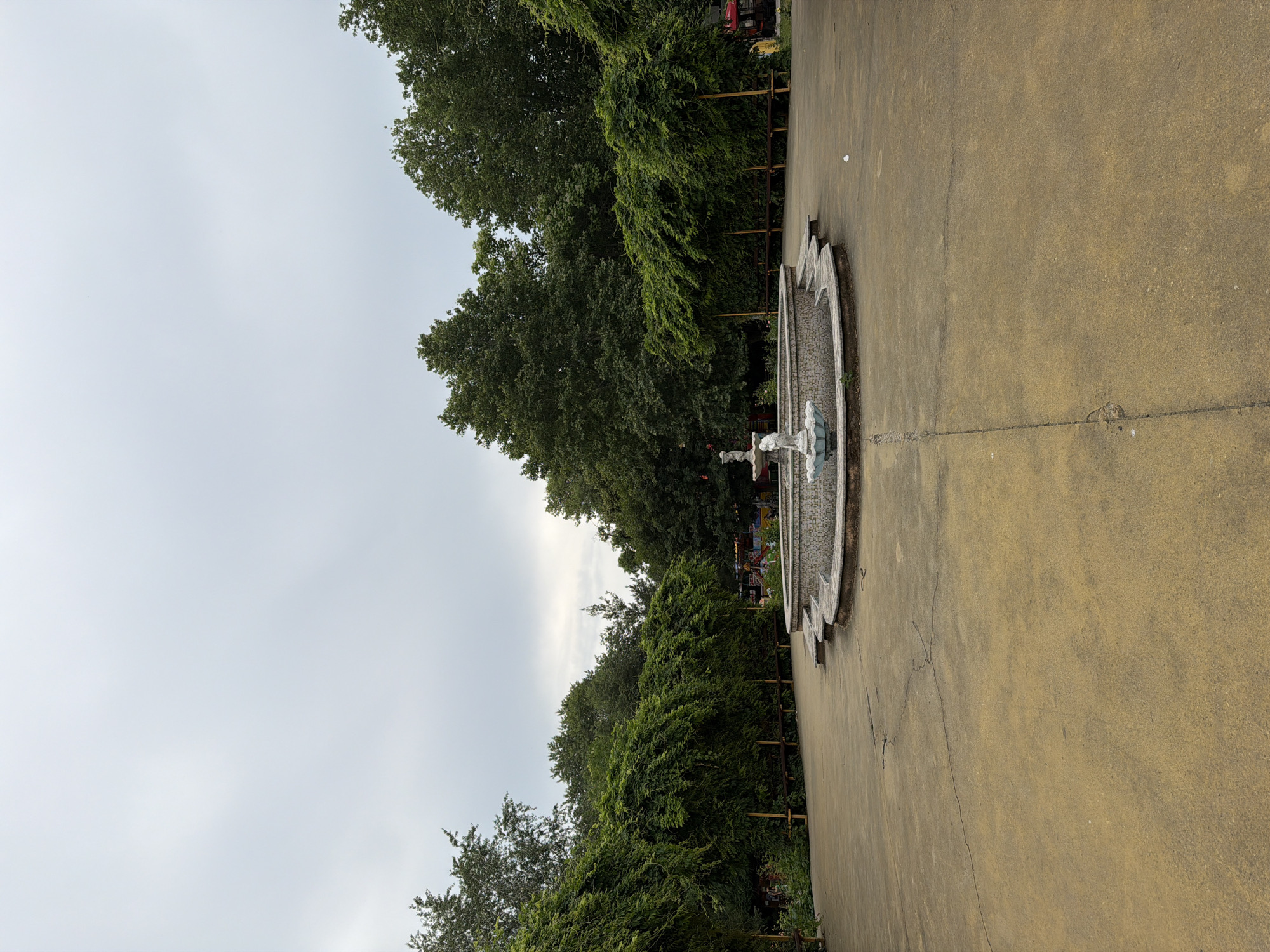 A circular, tiled fountain with a statue of a child is centrally located in an open, paved area. The fountain is surrounded by lush green foliage and trees, creating a park-like setting. The ground is a yellowish-brown color, with some visible cracks and debris. The sky is overcast. This scene is located in the Ivan Vazov City Garden in Pazardzhik, Bulgaria. There are no people visible in the image.