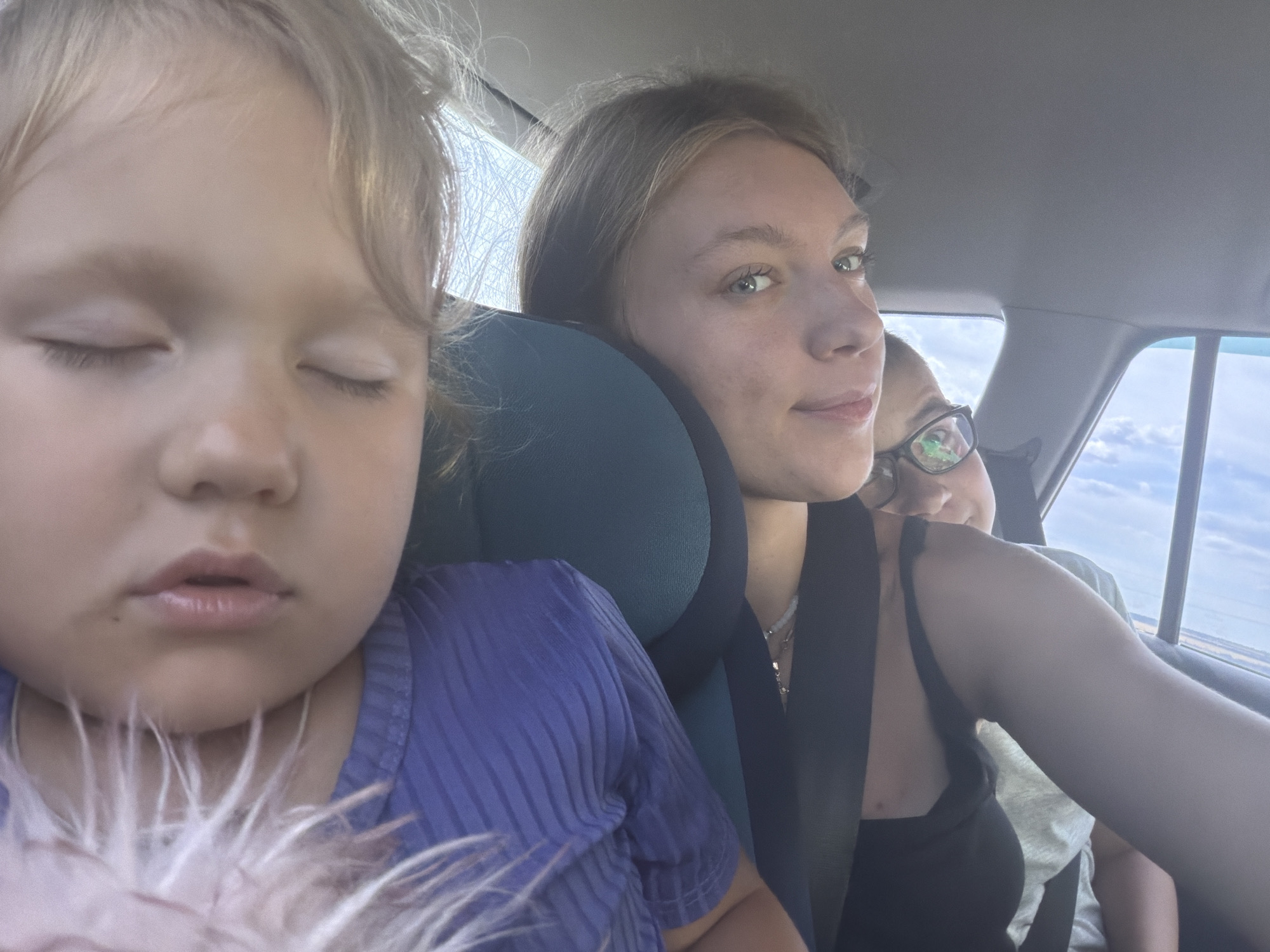 A close-up selfie taken inside a vehicle shows three people. In the foreground, a young child with closed eyes and blonde hair is wearing a purple ribbed shirt. To the right of the child, a young woman with blonde hair is looking at the camera. Behind her, another person wearing glasses is partially visible. The background shows the interior of a car with a headrest and a window with a view of a cloudy sky. The location is identified as Novoe Feizullovo, Russia.