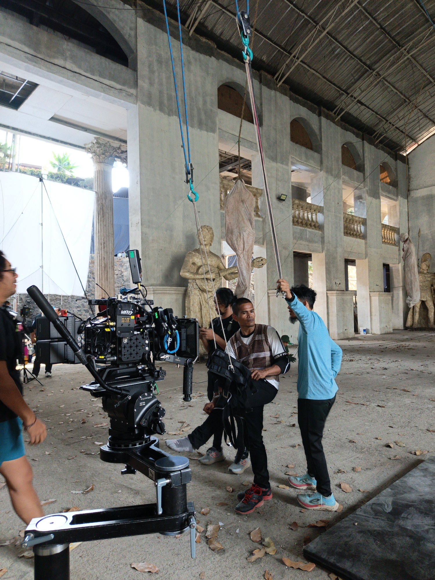 This image captures a film set within a derelict building in Cibogo, Indonesia. The scene is dominated by a large, complex film camera mounted on a professional tripod. Several individuals are engaged in production activities. One person, wearing a blue long-sleeved shirt, is holding a rope that extends upwards, suggesting the use of rigging or lighting equipment. Another person, in a checkered vest, is holding a camera accessory. Two other individuals are partially visible in the background. The environment appears to be an unfinished or abandoned structure with large arched openings, concrete columns, and a metal roof. Statues and draped fabric figures are present, adding to a theatrical or artistic atmosphere. Scattered leaves and debris suggest the location is outdoors or has a permeable roof.
