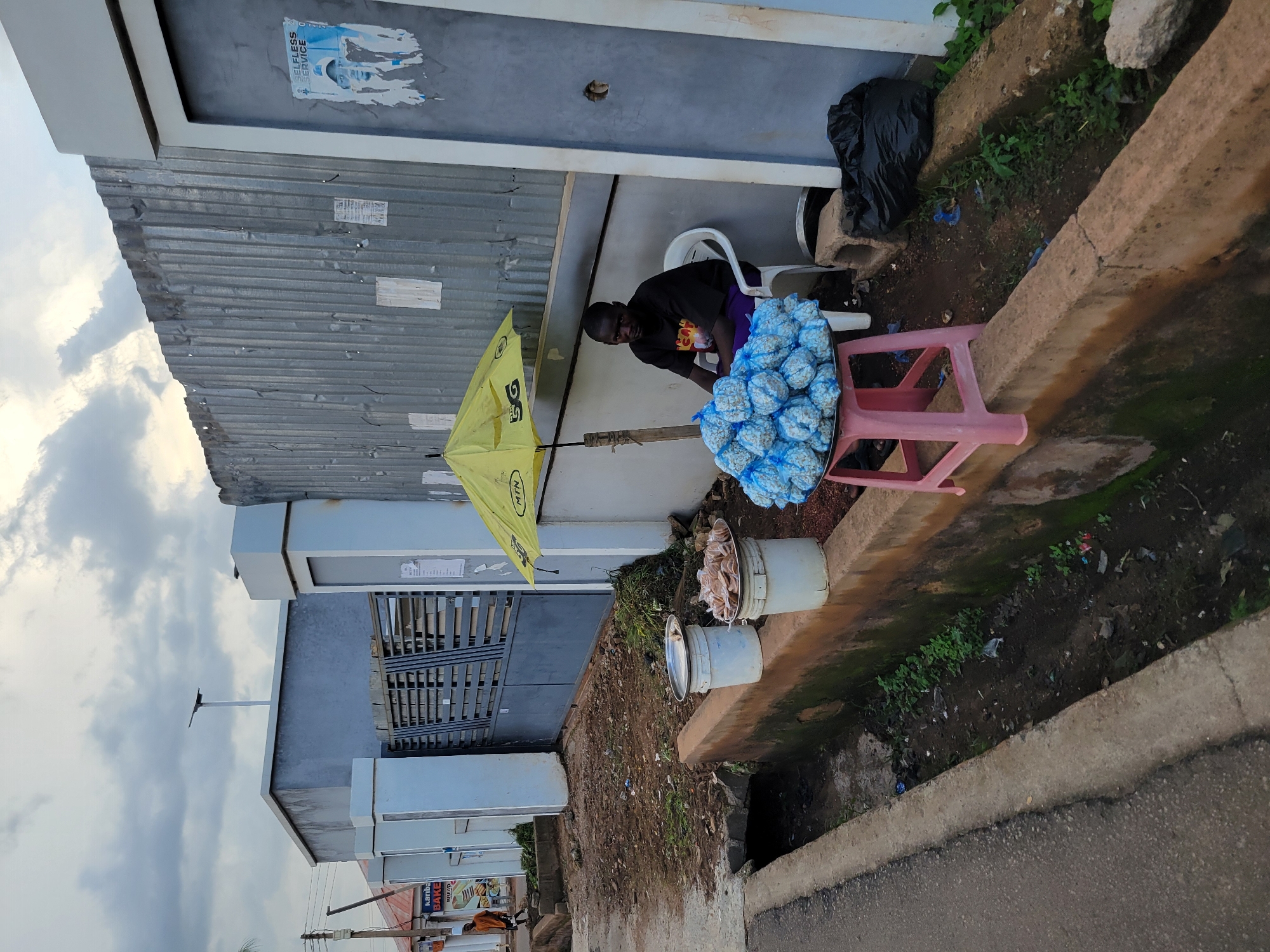 A man is sitting on a white chair next to a pink plastic stool, where a pile of what appear to be snacks wrapped in blue plastic are displayed. He is wearing a black t-shirt with a graphic design and purple shorts. A yellow umbrella with the "MTN" logo is open beside him, providing shade. Behind the man, a corrugated metal wall is visible, with a poster partially torn off. In front of the display, two white buckets are placed on the ground. One bucket is partially filled with food items wrapped in clear plastic. The scene appears to be outdoors, with a dirt path and a concrete drainage ditch to the right. The overall setting suggests a street-side market or vendor stall. The sky is cloudy, indicating overcast weather. The image has been rotated, so the subjects and scene are oriented sideways. There is text on the yellow umbrella that reads "MTN" and on a poster on the wall, though much of it is illegible.