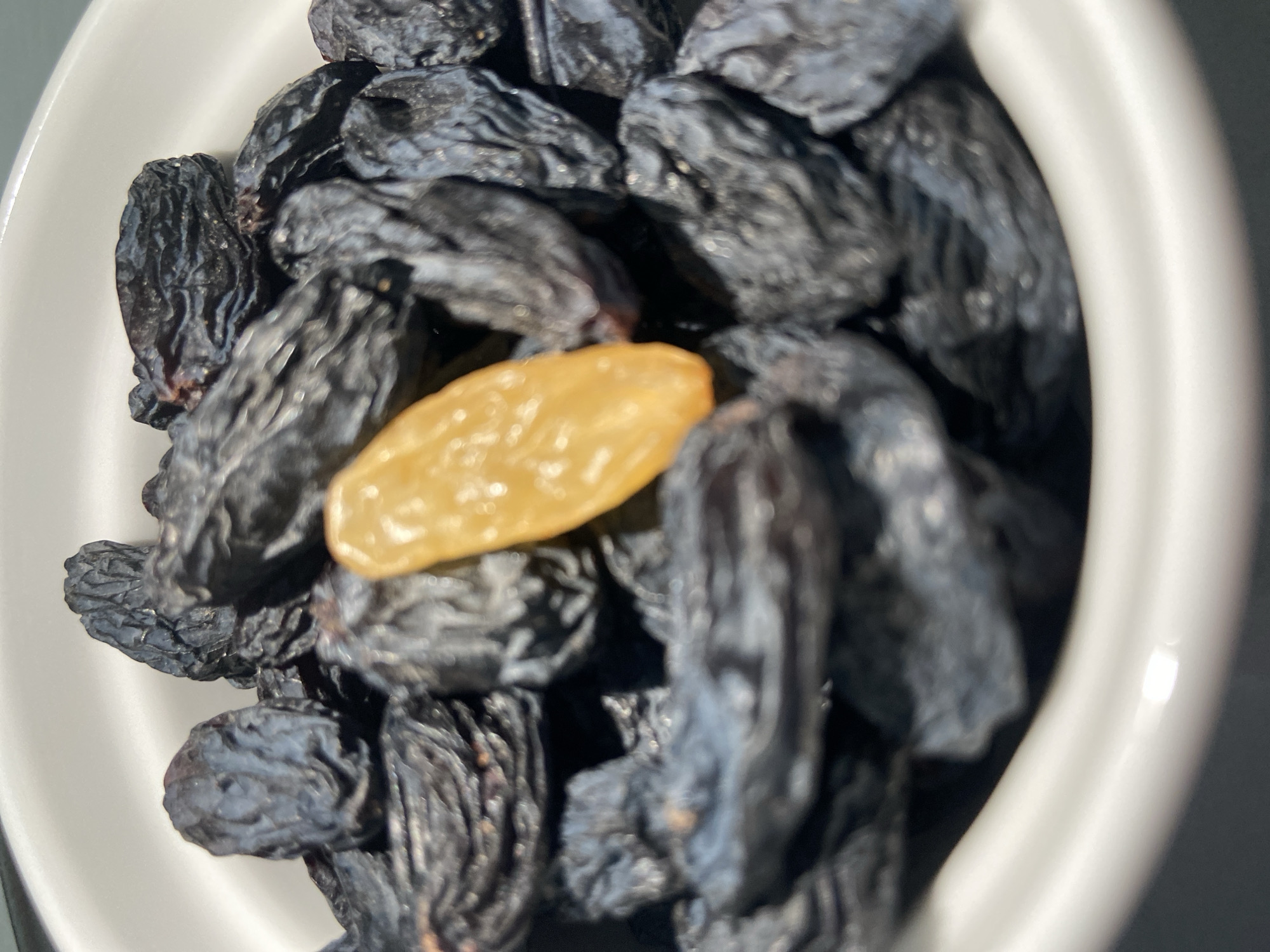 A close-up shot shows a white bowl filled with dark-colored raisins, among which a single golden raisin is centrally located. The lighting appears to be natural, possibly daylight. The bowl is partially visible, indicating it is the container for the raisins. The scene's location within Budva, Montenegro, is not discernible from the provided image.