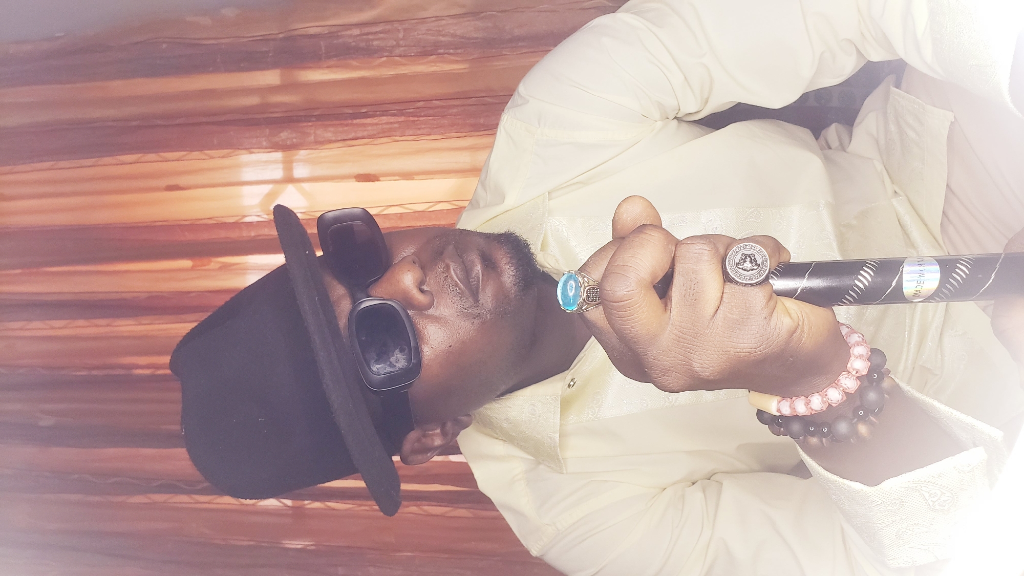 A man wearing a black fedora and sunglasses is angled in the center of the frame. He wears a light yellow, patterned, long-sleeved shirt with decorative rings on his right hand and bracelets on his wrist. He holds a black stick or cane in his hand. The background appears to be a patterned wall or curtain. It is impossible to confirm the location in Narayi, Nigeria, with the image alone.
