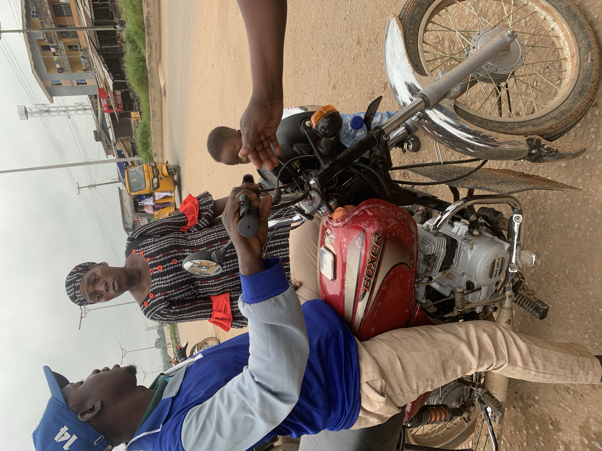 In Shagamu, Nigeria, a medium shot captures a man in a blue jacket and beige pants seated on a red motorcycle, facing left and engaged in conversation. A woman in a black and red patterned dress stands behind him, also facing left. Between the man and the motorcycle's handlebars, a young child looks towards an outstretched hand. The background reveals a street scene with buildings, a yellow motorized rickshaw, and power lines under an overcast sky. The ground is a dirt and gravel surface, indicating an outdoor, possibly roadside, setting.